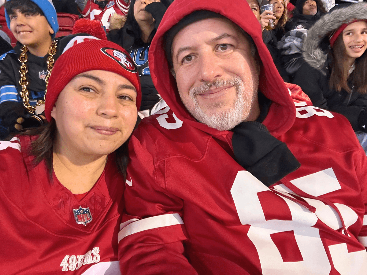 A couple at a football stadium taking a selfie, both wearing red San Francisco 49ers jerseys and hats, smiling with fans in the background.