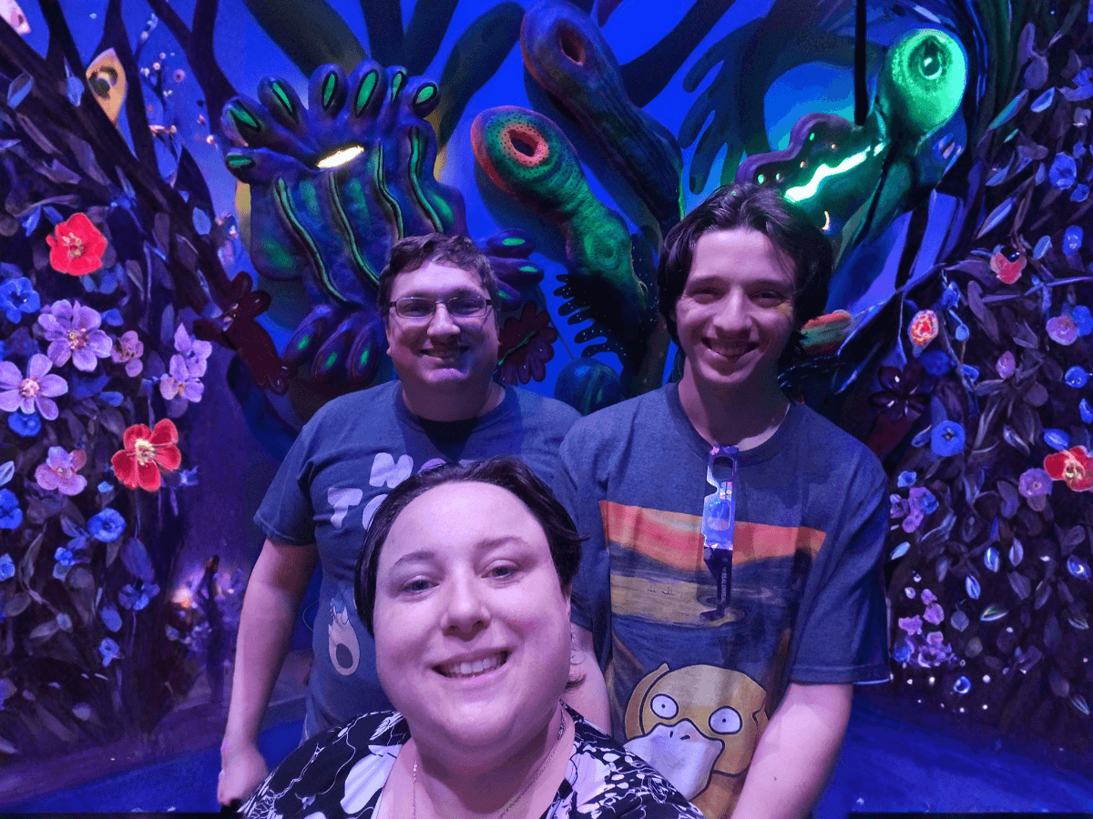 Three friends smile for a selfie in a neon blue underwater exhibit with colorful coral and fantastical plants behind them.