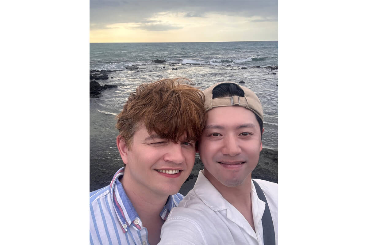 Two friends taking a selfie on a rocky beach at sunset, with waves and a cloudy sky in the background.