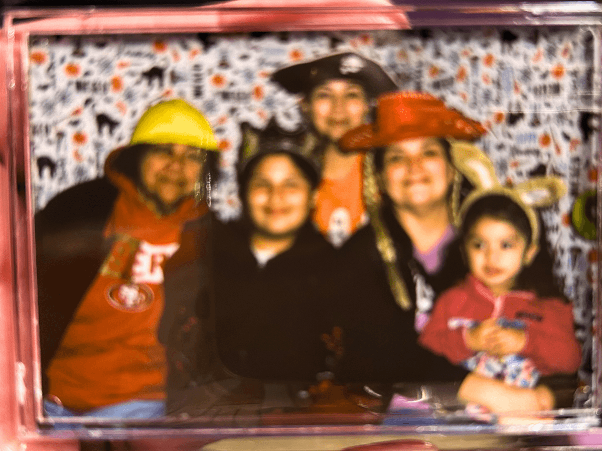 Group of five people posing in a photo booth, wearing colorful, novelty hats (yellow helmet, red wide-brimmed hat, etc.) in front of a patterned backdrop.