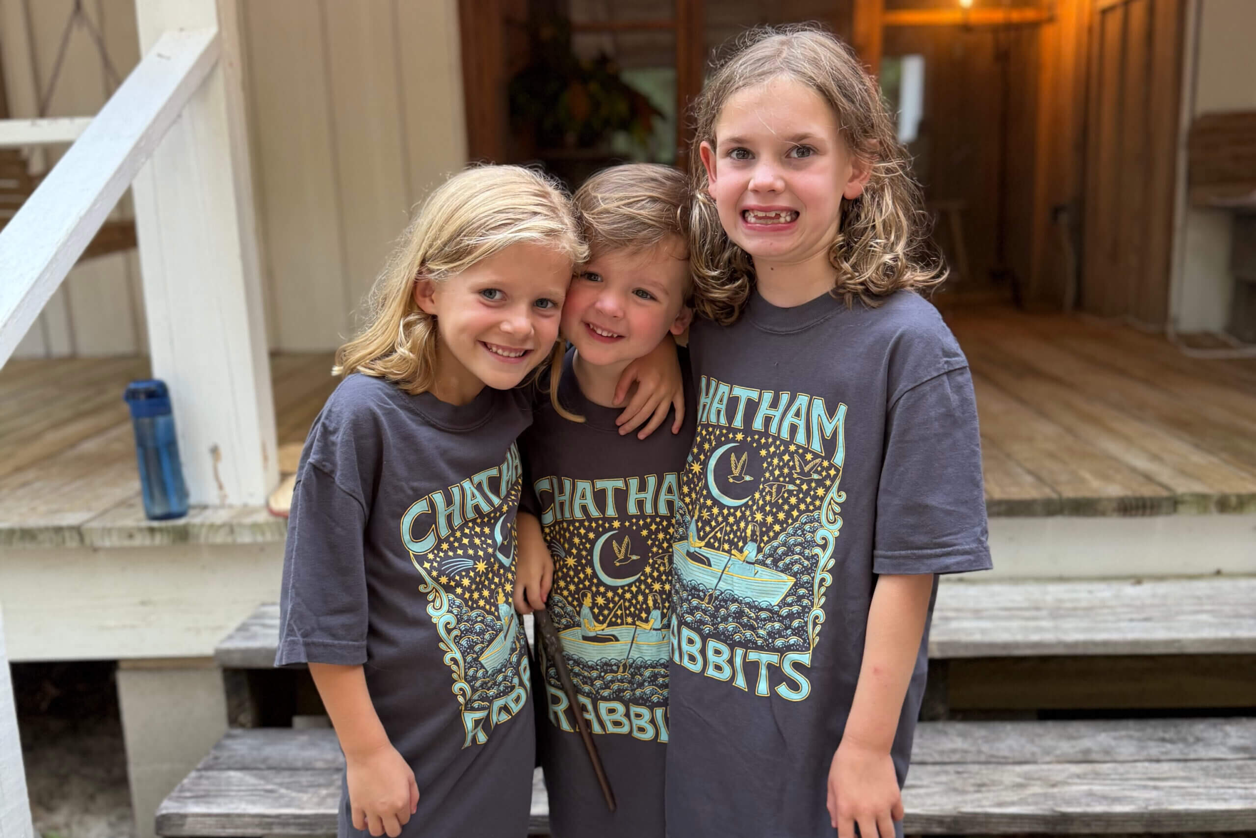Three smiling children in matching dark gray shirts hug on wooden steps outside a house.