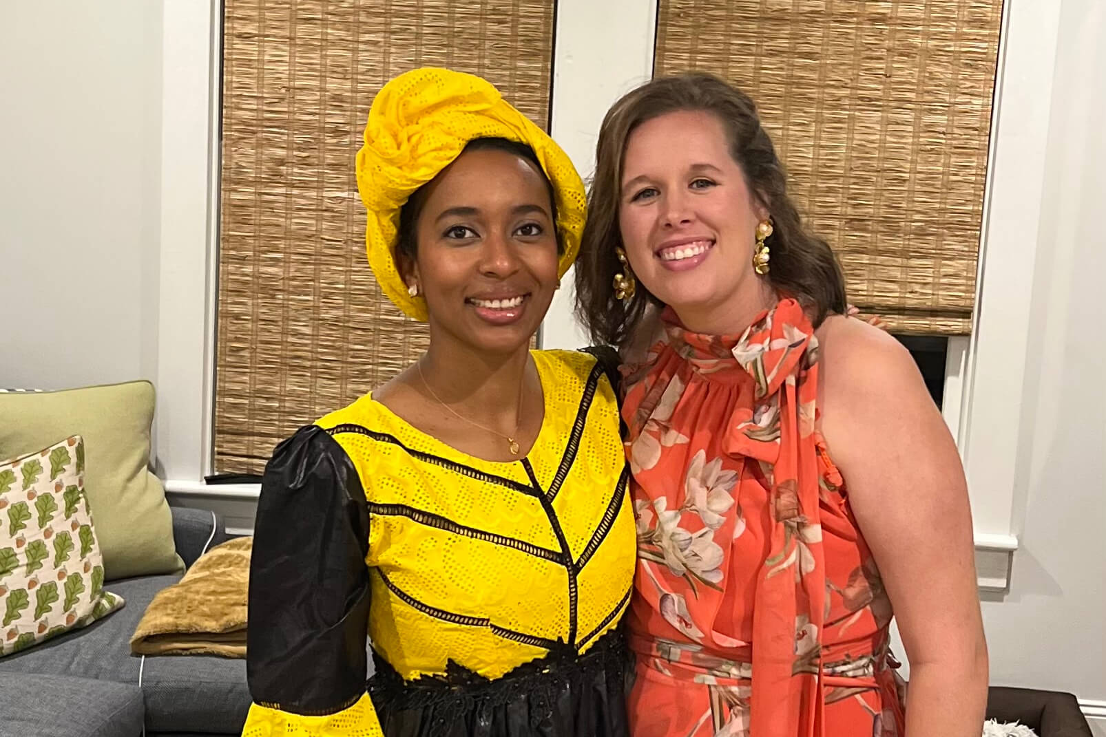Two women smiling indoors; one in a bright yellow traditional dress and head wrap, the other in an orange floral dress.