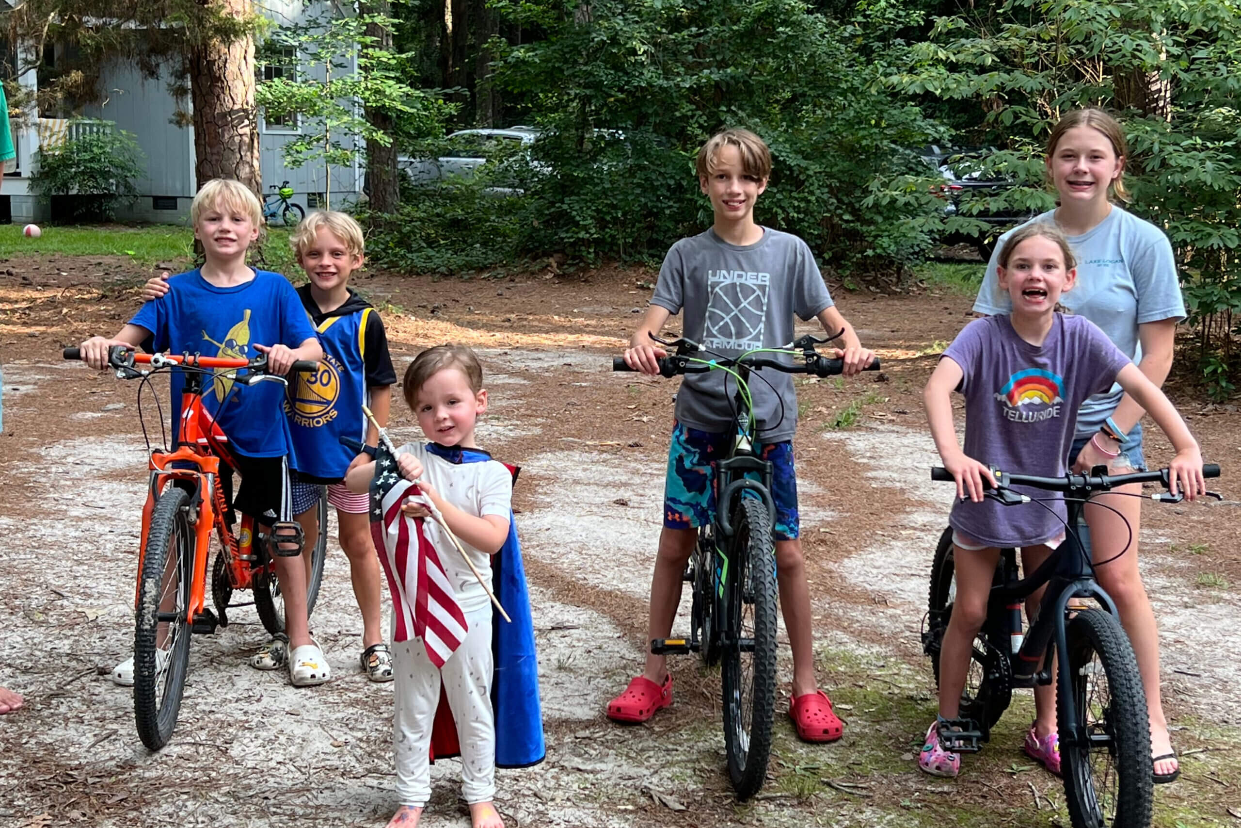 Six kids with bicycles posing on a dirt path in a wooded backyard, smiling at the camera.