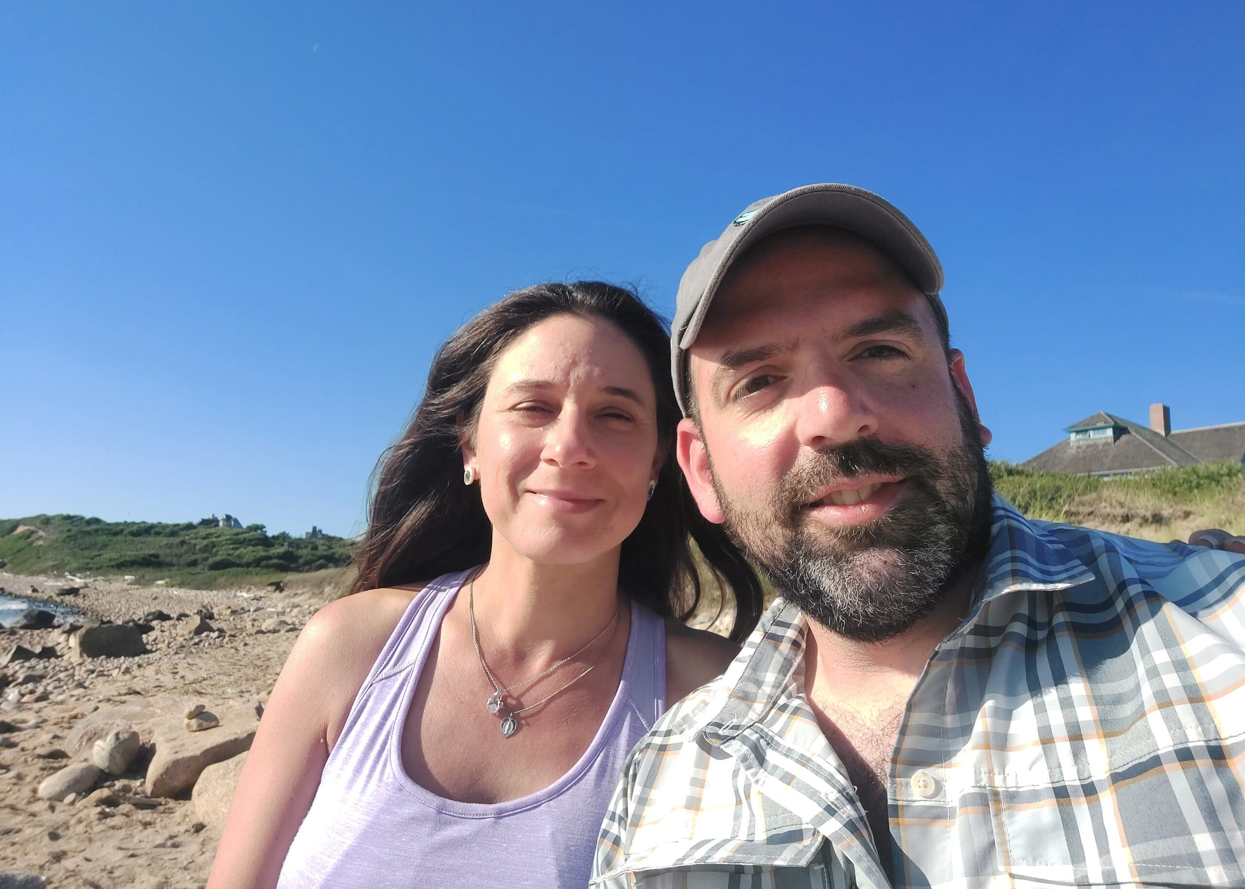 Smiling couple taking a beach selfie on a sunny day with a rocky shore and clear blue sky behind them.