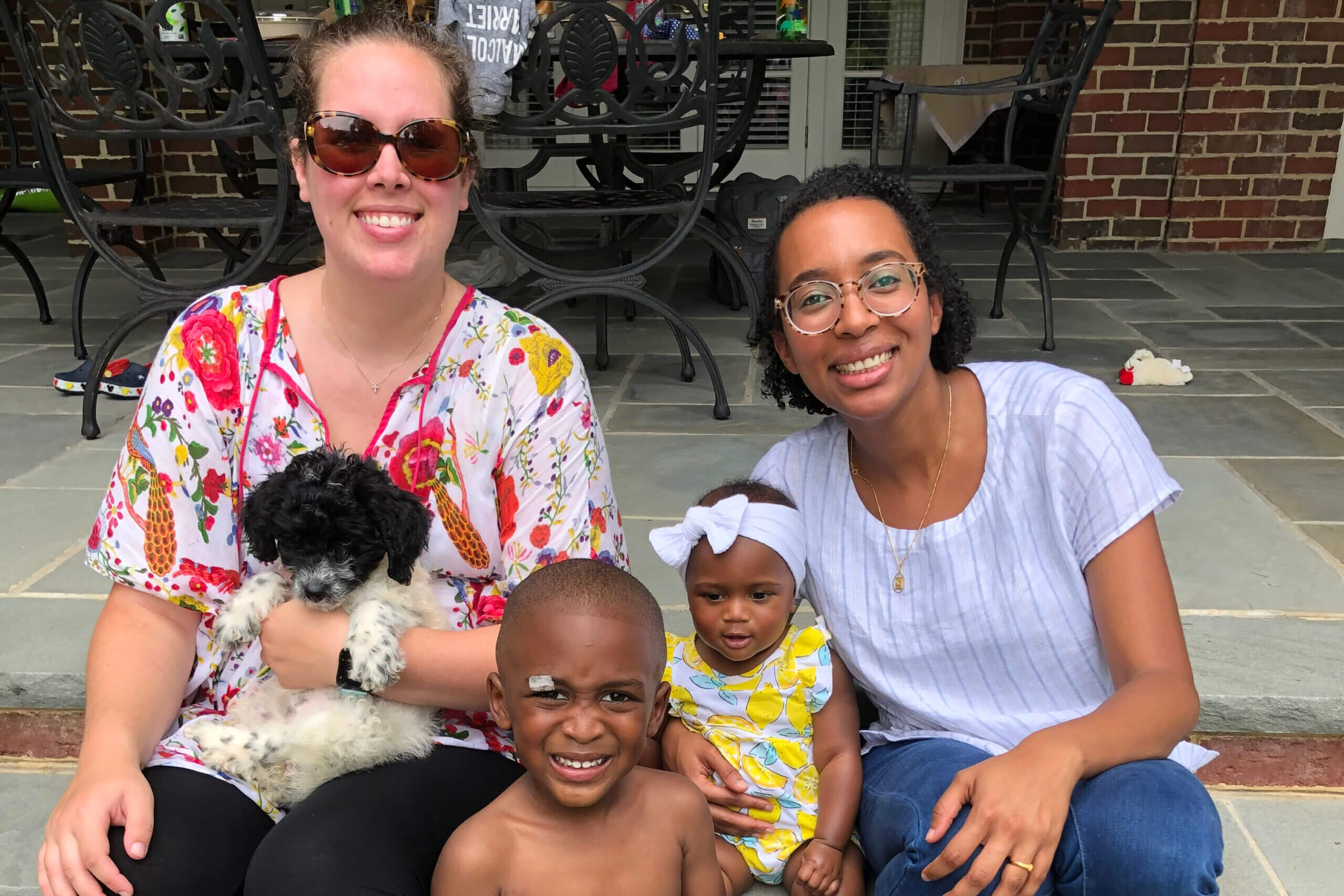 Smiling group of two women and three children sitting on a brick porch with a small white fluffy dog on a lap in the foreground.