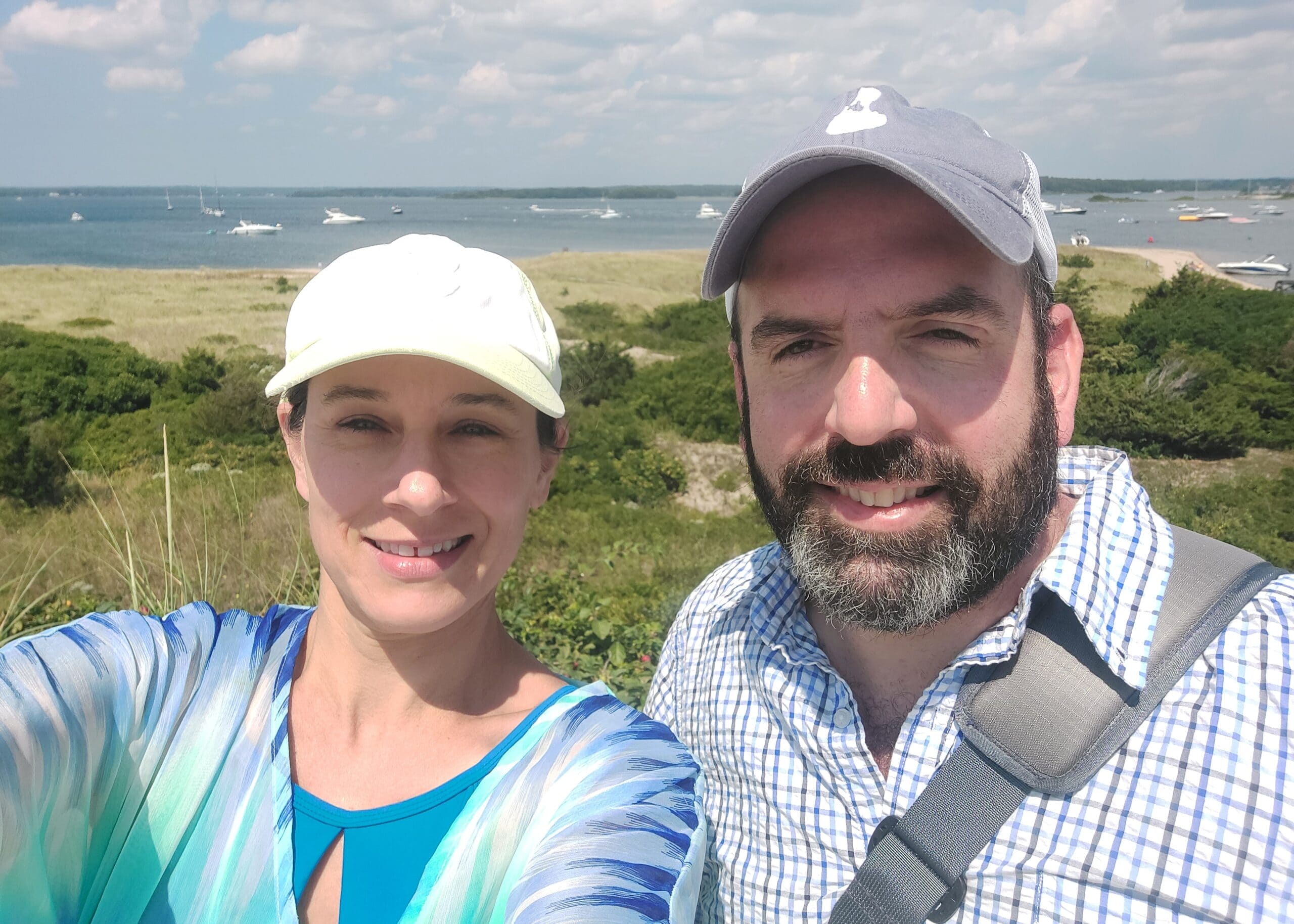 Smiling couple taking a beach selfie with boats on the bay in the background.