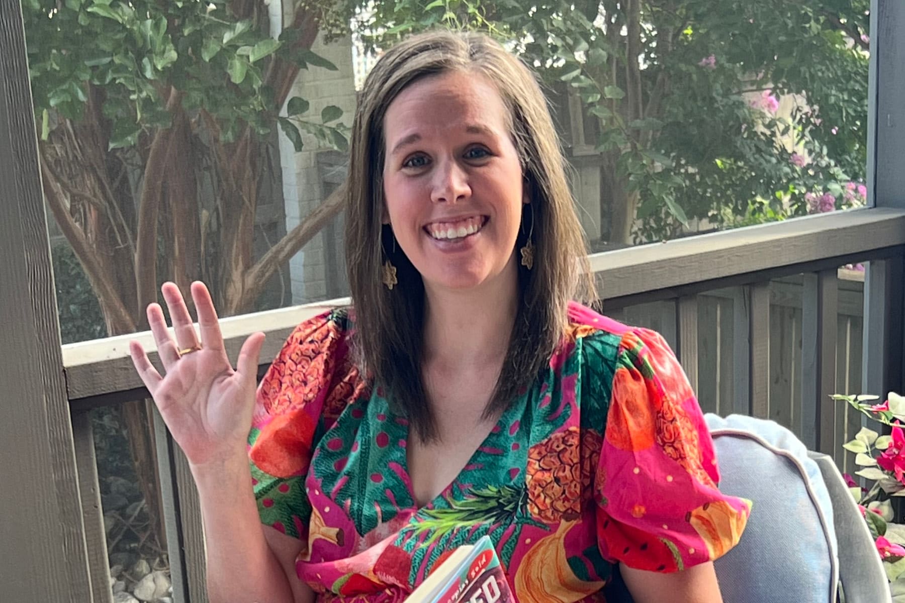 Smiling woman with shoulder-length hair sits on a porch, wearing a bright tropical dress and waving at the camera while holding a colorful magazine.