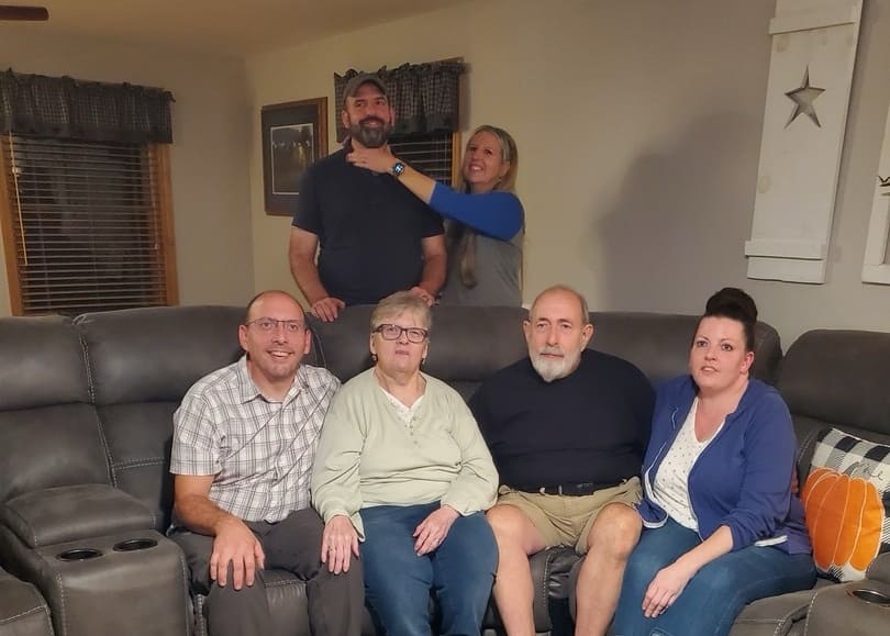 Group of six adults posing on a living-room sofa, with two standing in back and four seated front, smiling at the camera.