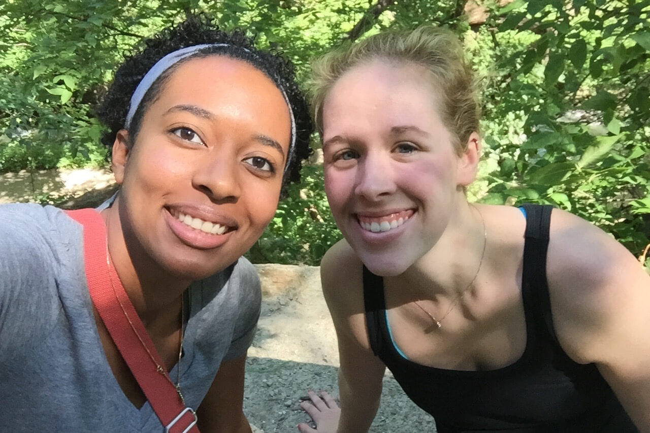 Two friends taking a selfie outdoors, smiling with green trees behind them.