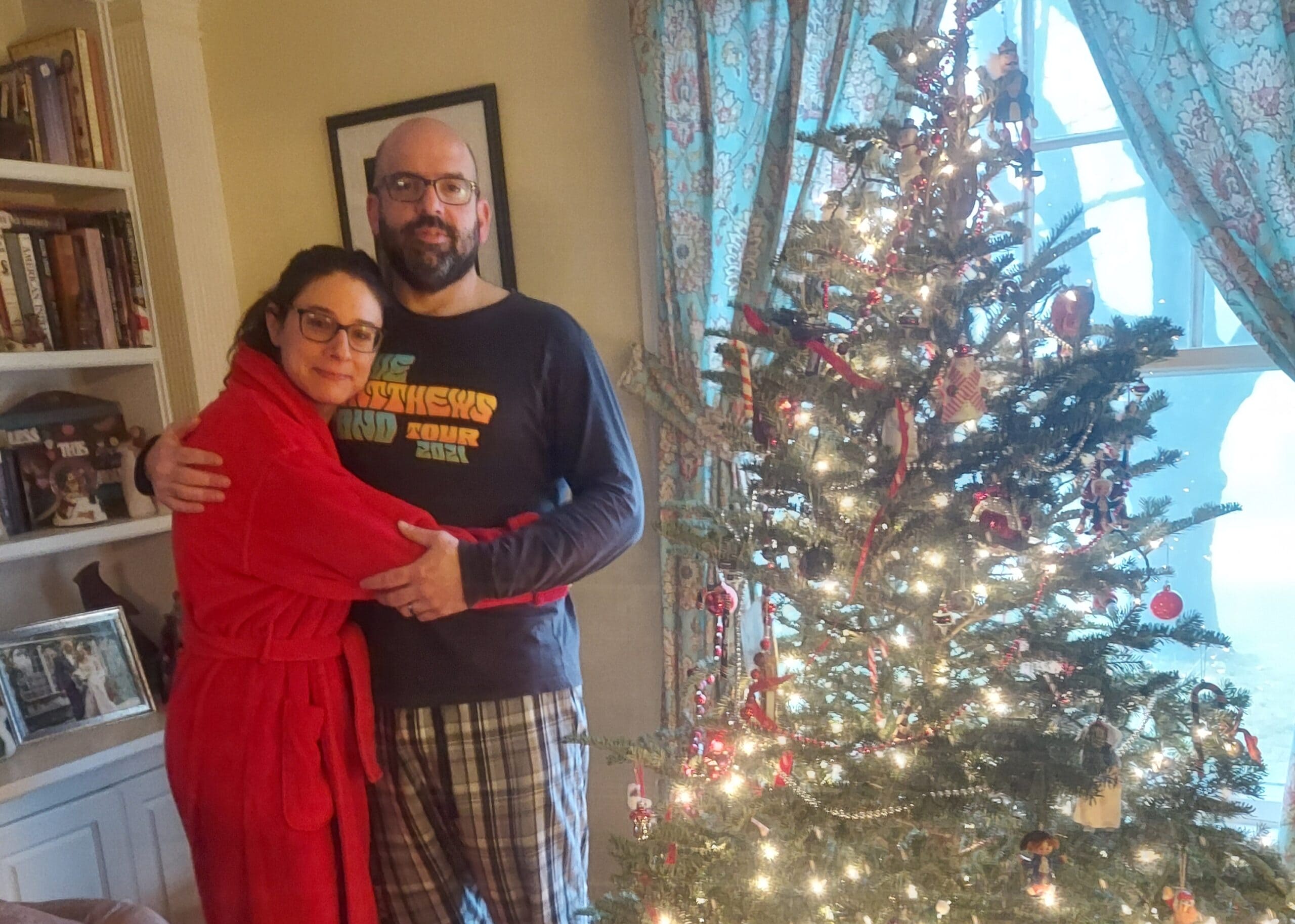 A couple embraces beside a decorated Christmas tree in a living room setting.