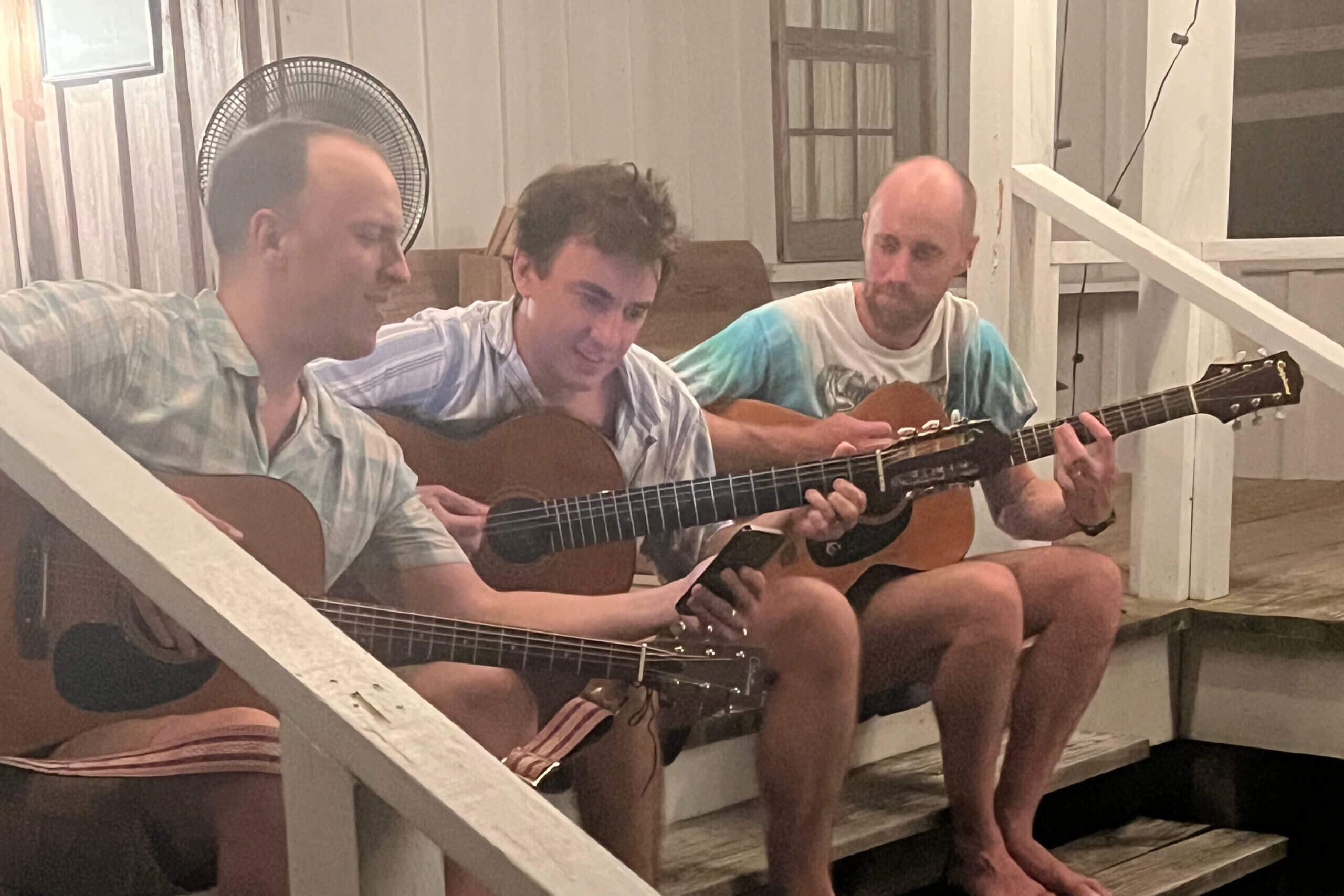 Three men sit on wooden stairs indoors, playing acoustic guitars together in a casual jam session.