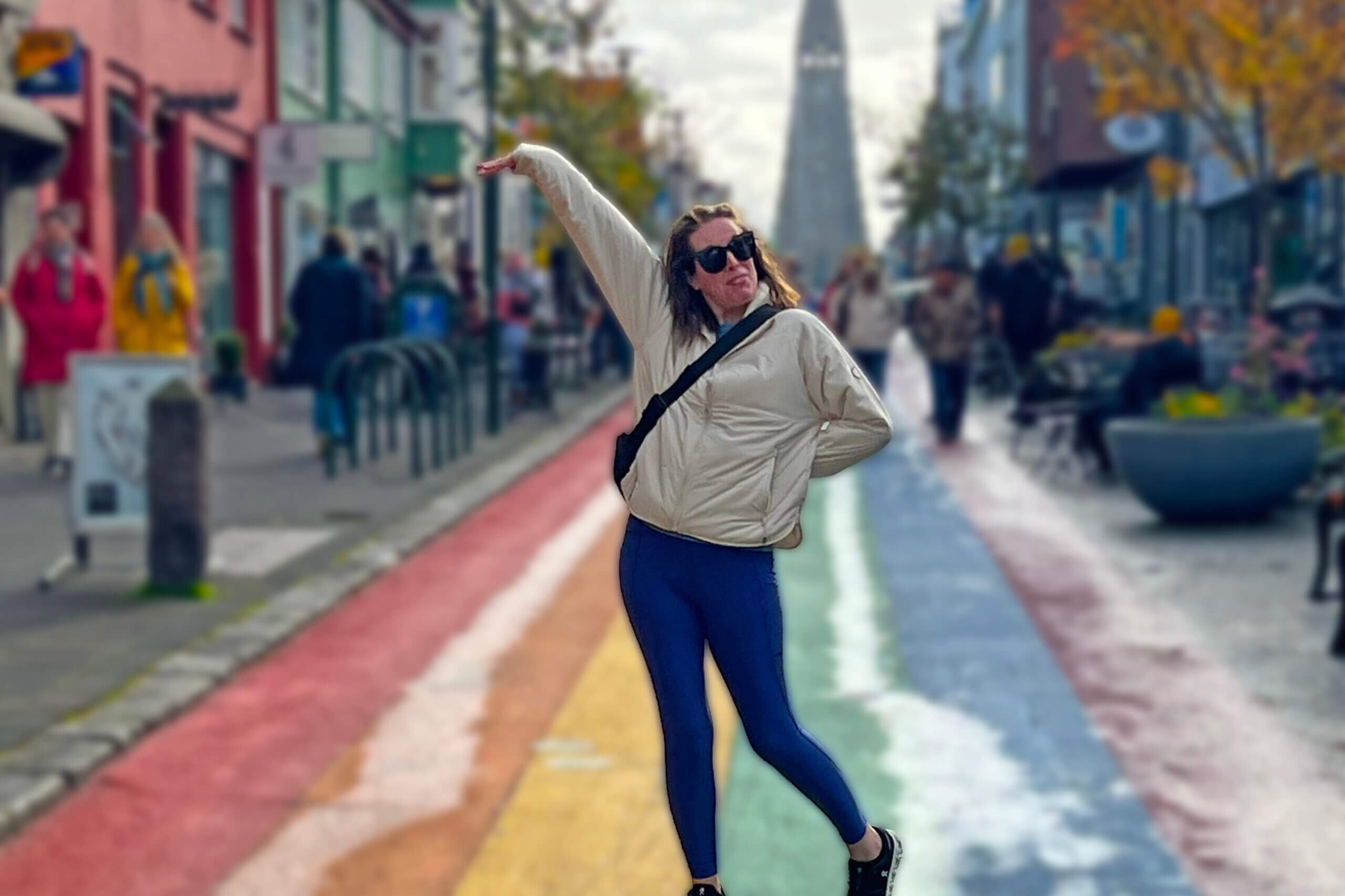 Woman in a beige jacket and sunglasses dancing on a rainbow-colored street with shops behind her.