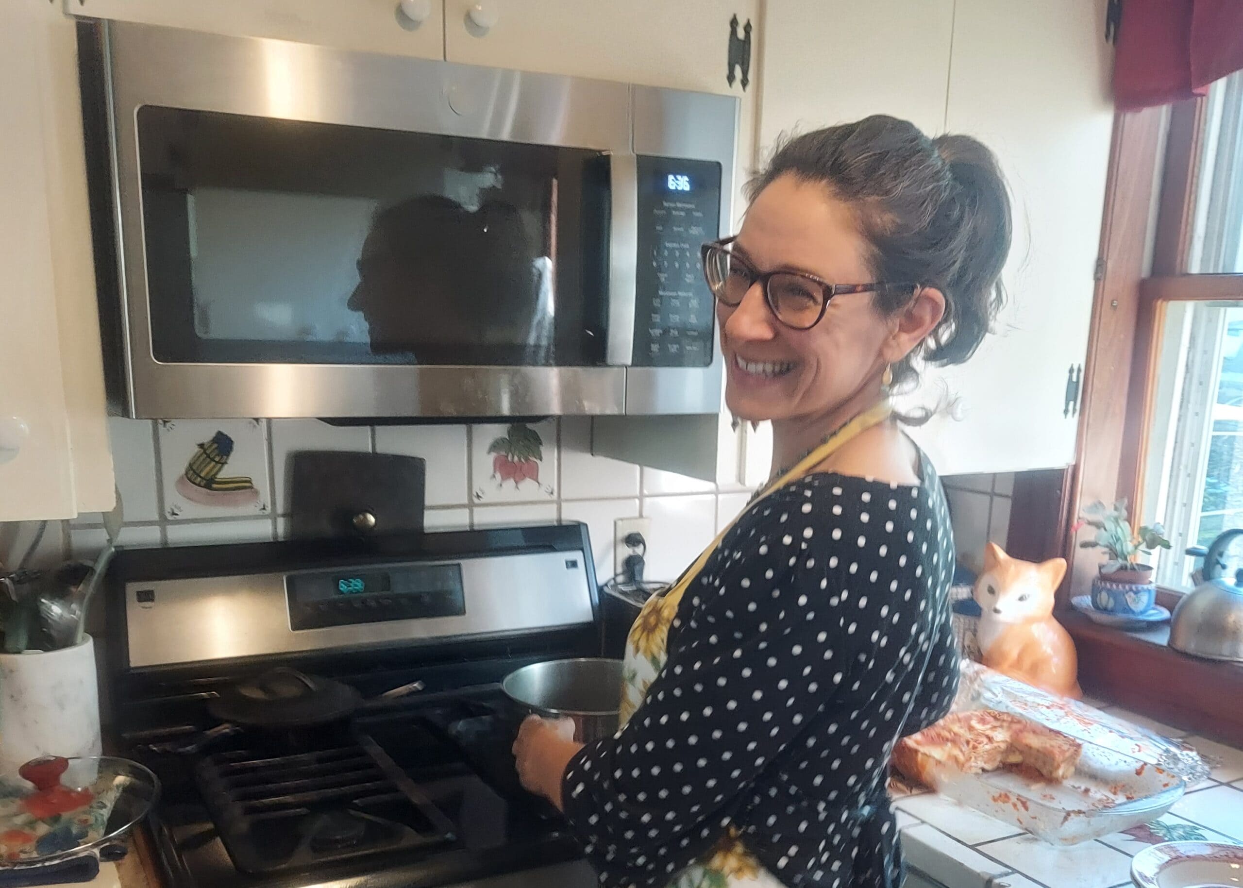 Woman in a patterned dress and apron smiling in a kitchen, standing by a stainless steel microwave and stove.
