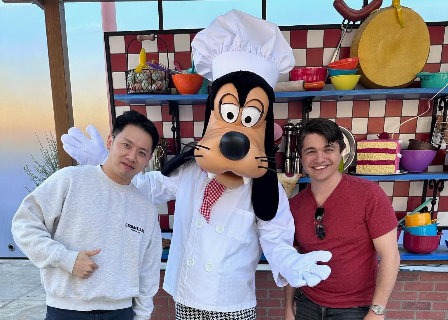 Three people posing with a large cartoon dog-costumed character in a chef's outfit and hat in a colorful kitchen set with bowls and utensils behind them.