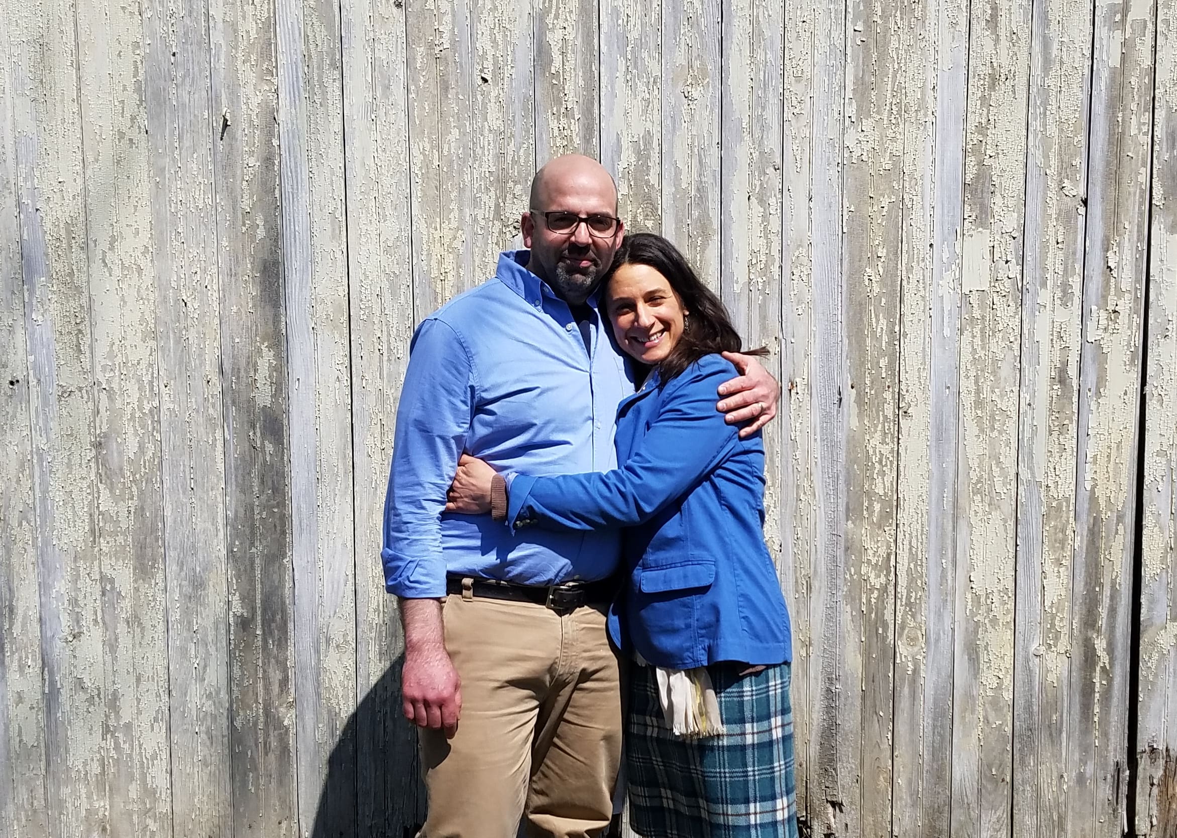 Couple wearing blue shirts embrace each other against a weathered wooden wall.