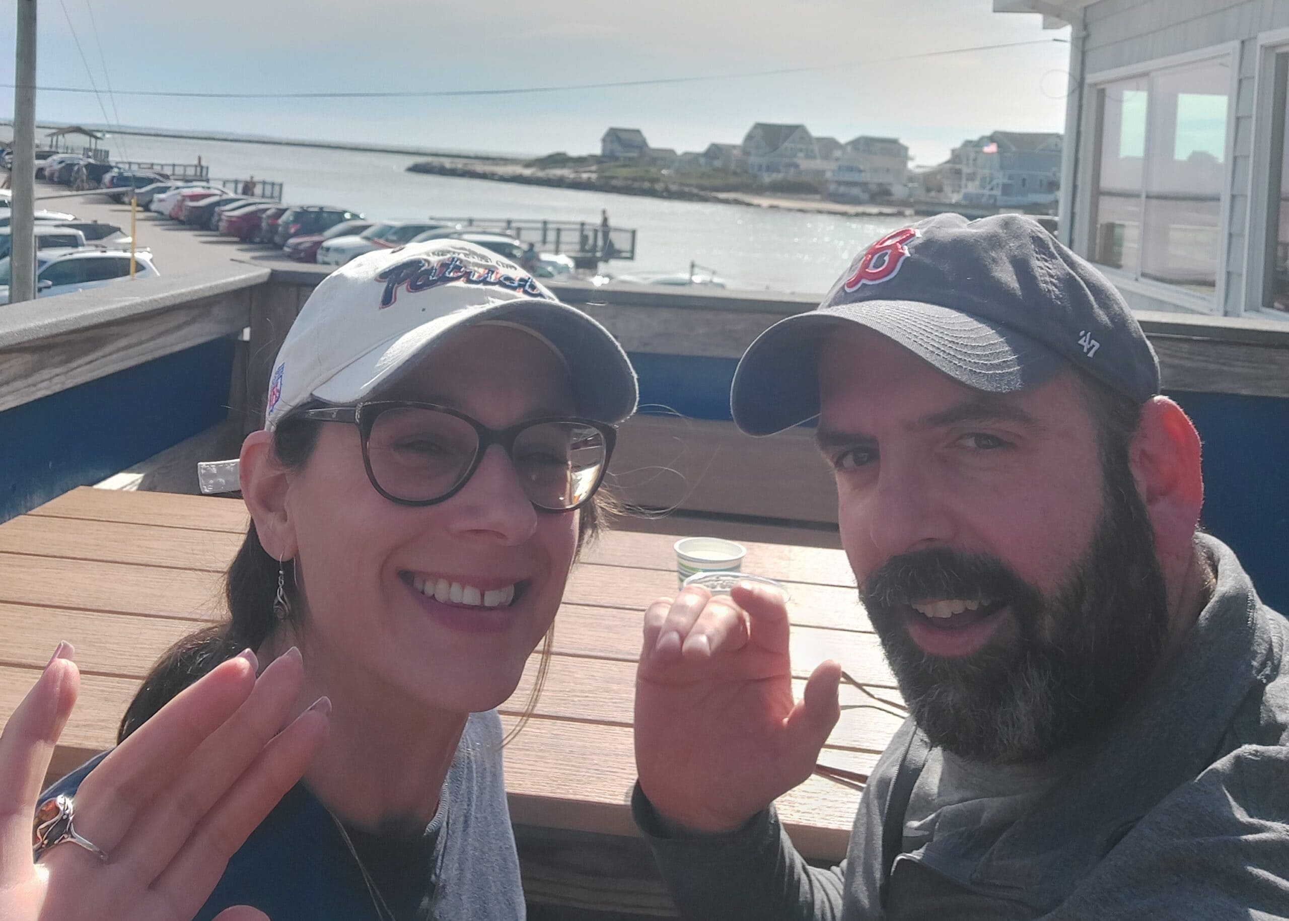 Selfie of a smiling couple at a seaside deck with a marina and houses in the background, both wearing caps.