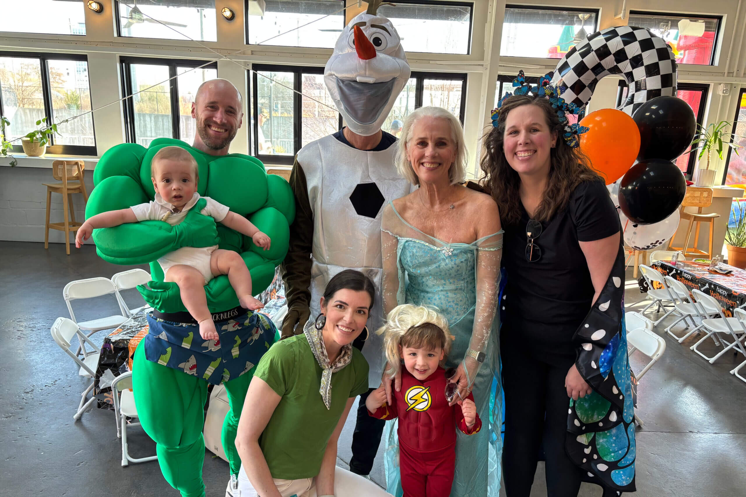 Group of adults and children in costume posing for a photo at a party, including a baby in a Hulk suit, an Olaf character, Elsa, and others with balloons in the background.