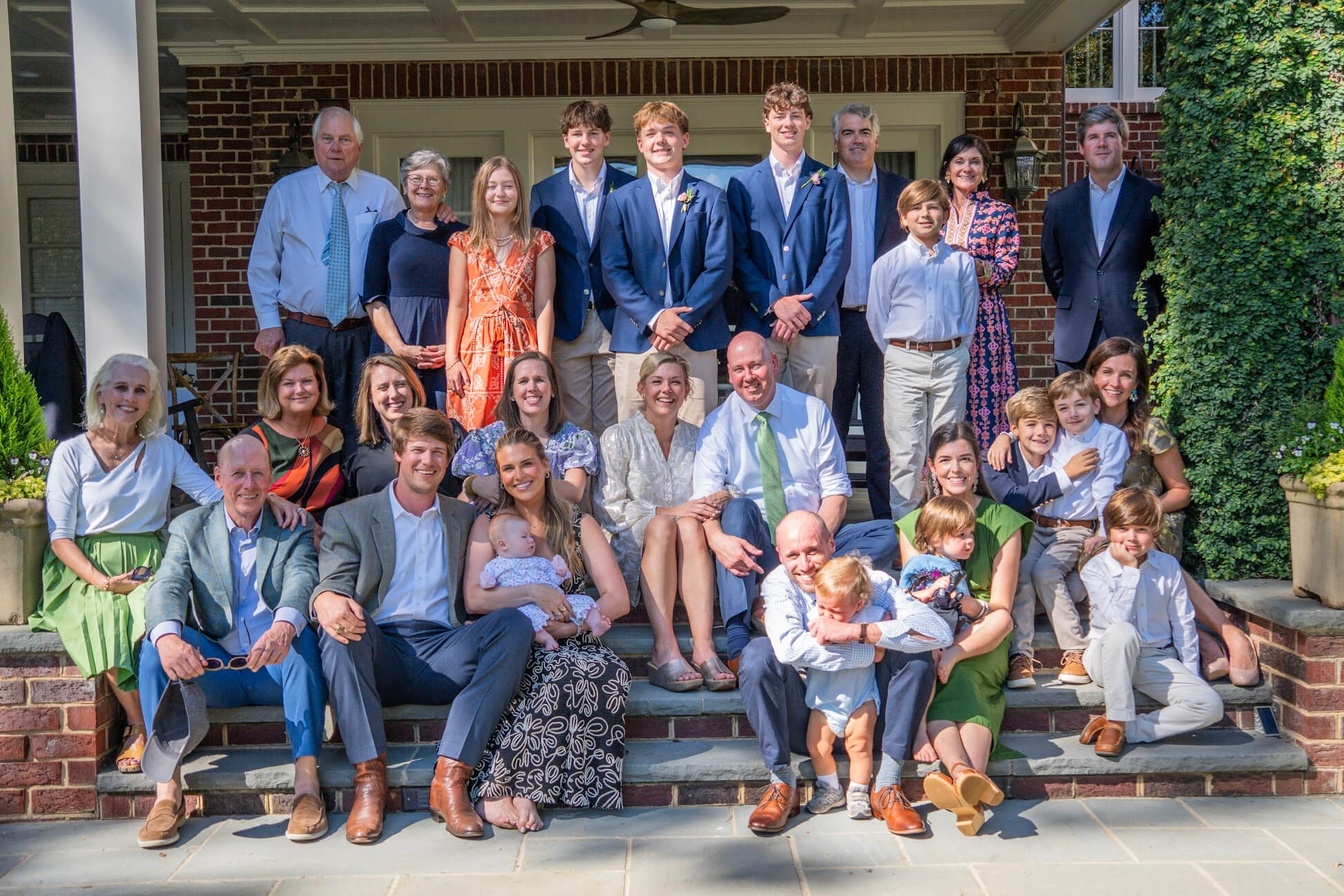 Large multi-generational family group photo on brick house steps, with adults, teens, and children in smart casual and formal outfits.