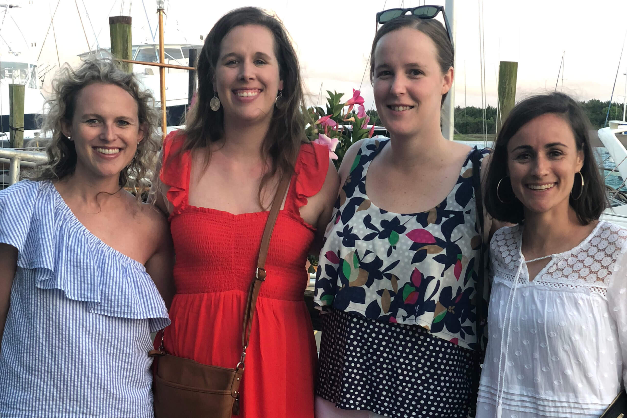 Four women smiling and posing together at a marina with boats in the background, in summer outfits.