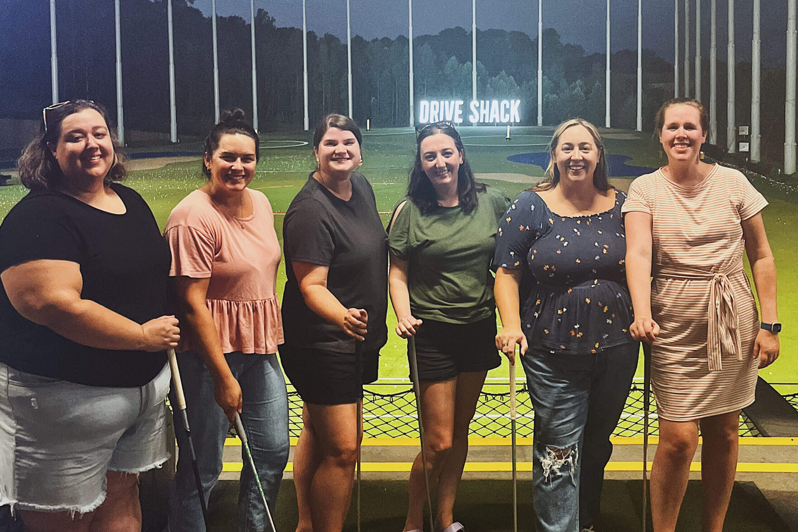 Six women standing side by side indoors at a Drive Shack venue, smiling and holding golf clubs.