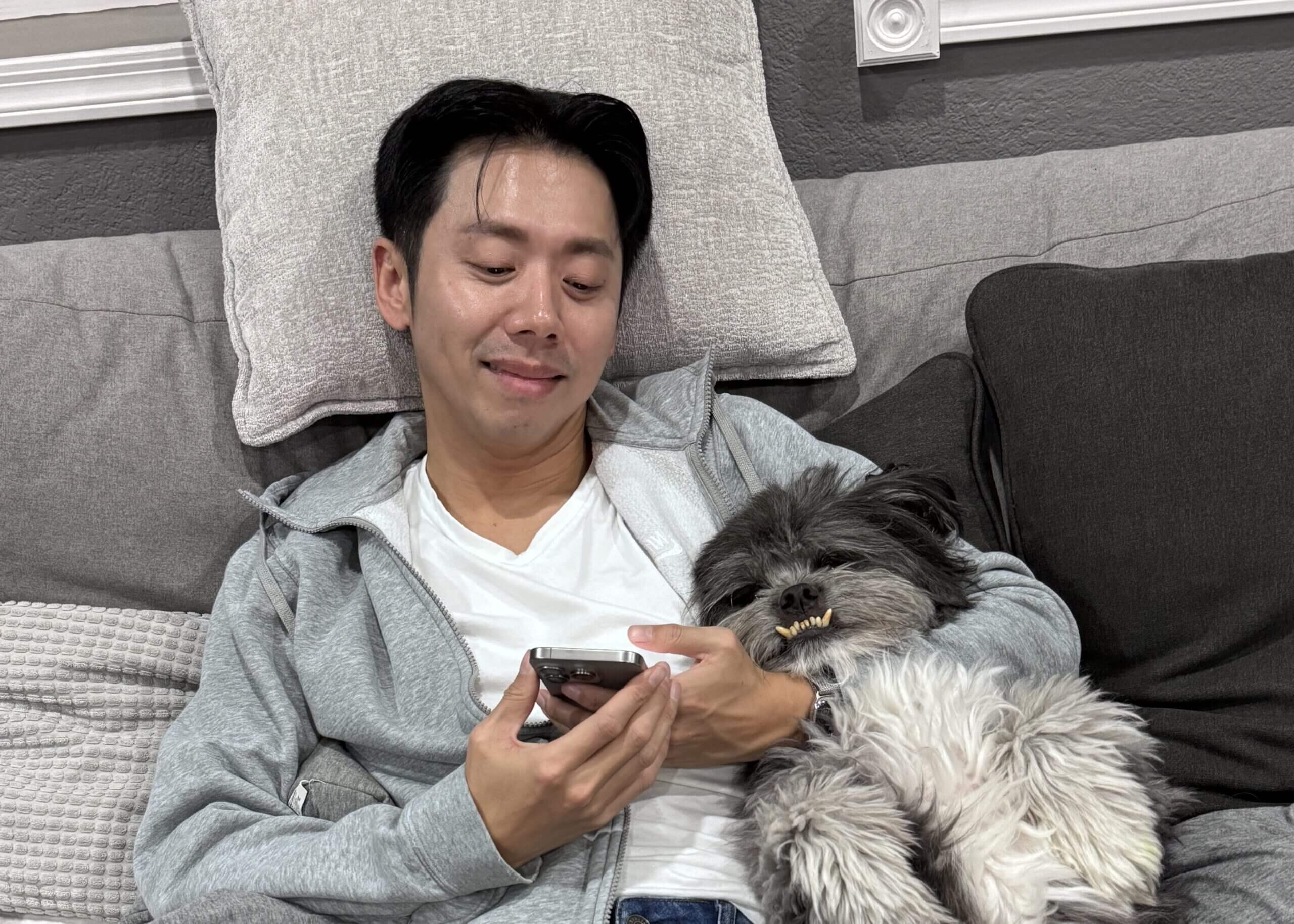 Man in a gray hoodie lying on a couch, looking at a smartphone while a fluffy dog snoozes against his chest on his lap.
