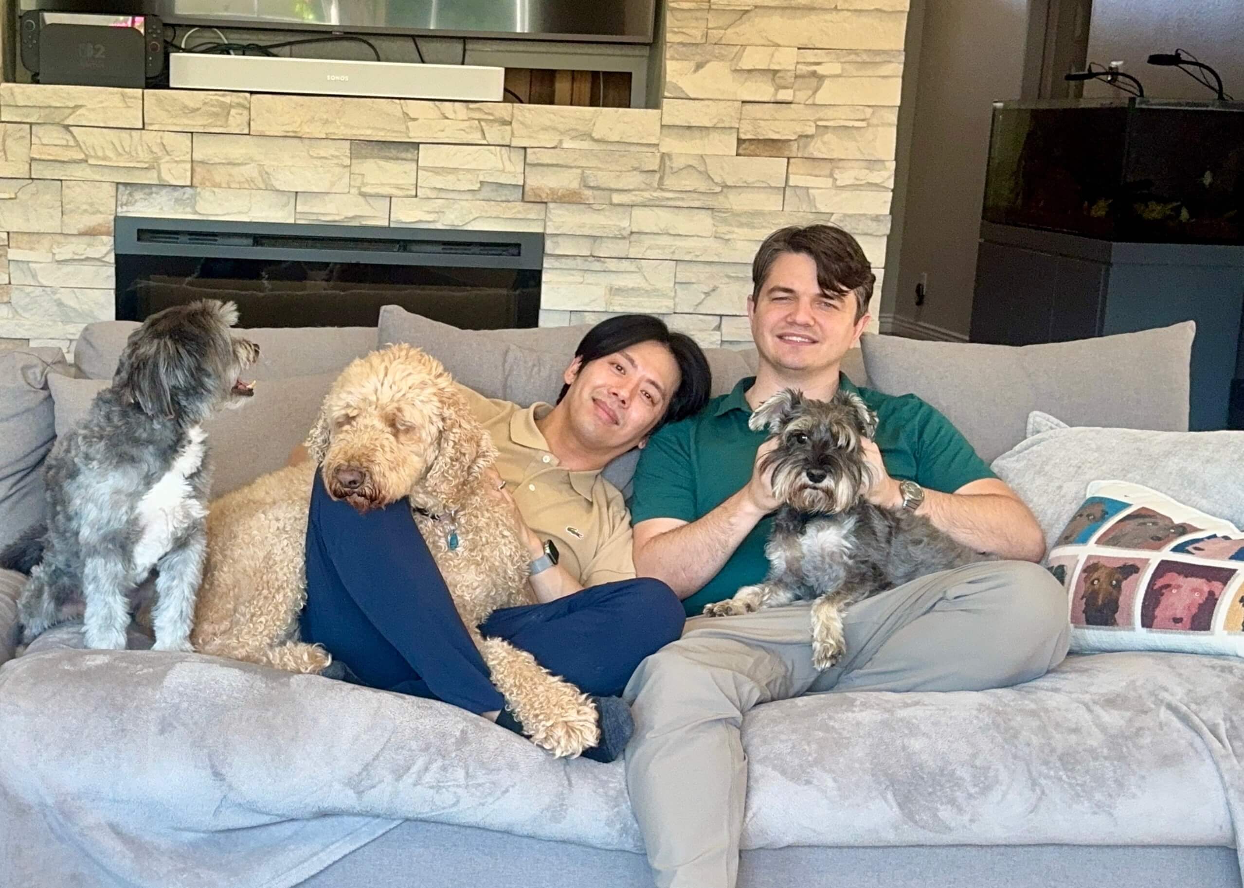 Two men sit close on a gray sofa in a living room with a stone fireplace; they hold three dogs—a gray-and-white terrier on the left, a curly beige poodle mix in the middle, and a Schnauzer on the man's lap on the right.