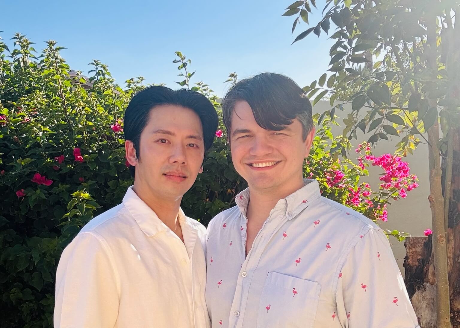 Two men stand side by side outdoors, smiling, with green bushes and pink flowers behind them under a sunny blue sky.