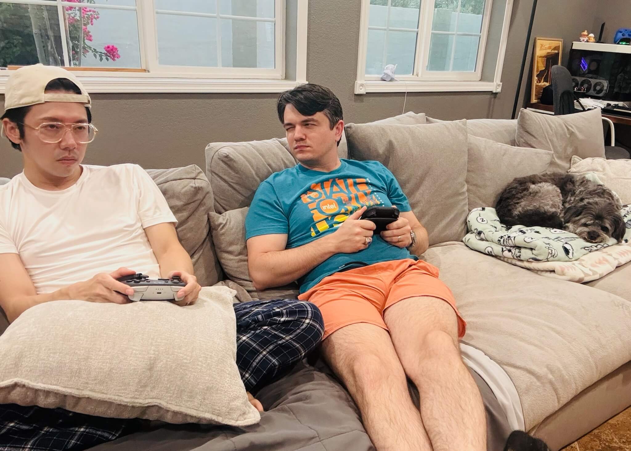 Two people sit on a beige couch holding game controllers, with a small dog sleeping on a blanket nearby in a living room.