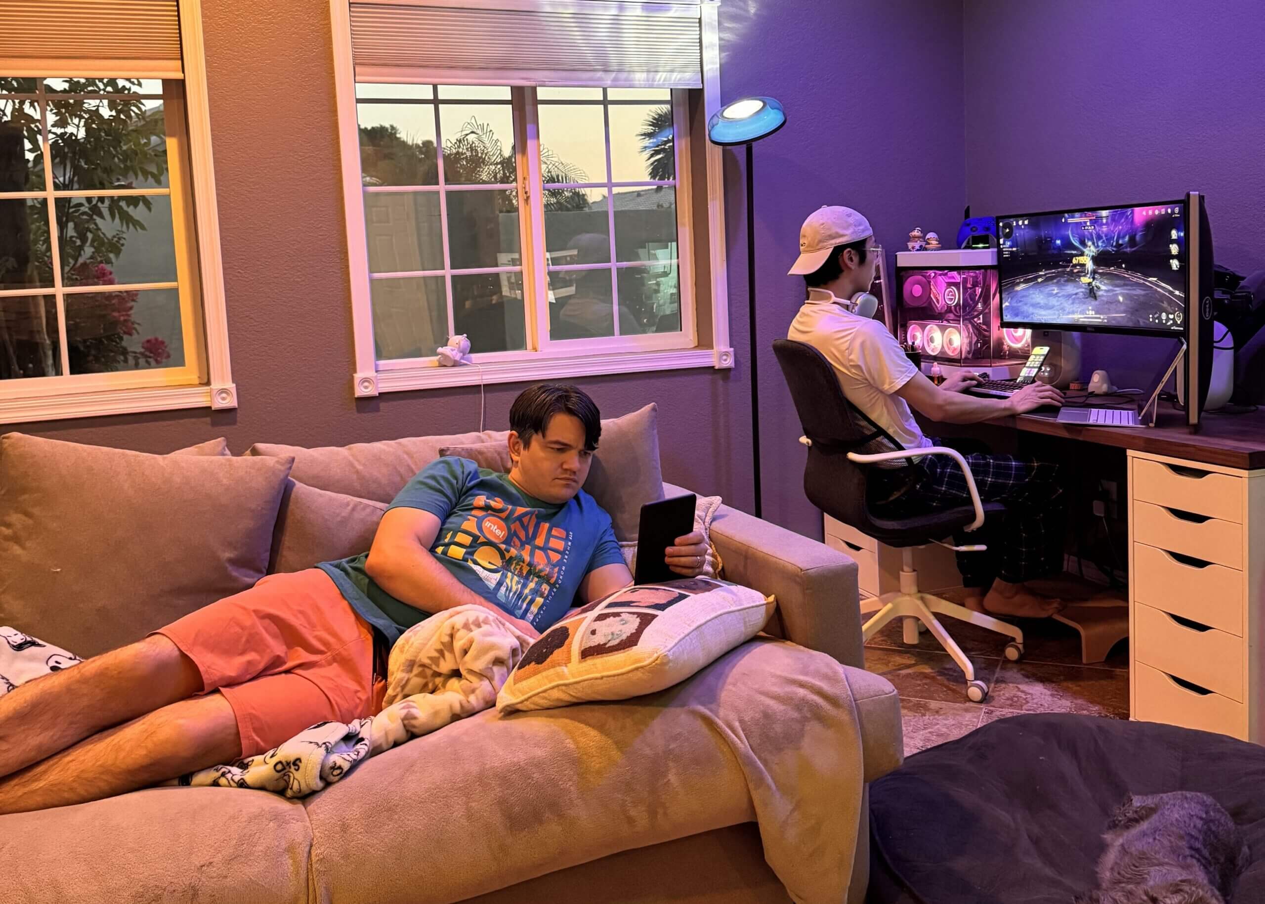Person on a beige couch using a tablet while another gamer sits at a desk with a large monitor and purple lighting in a home setup.
