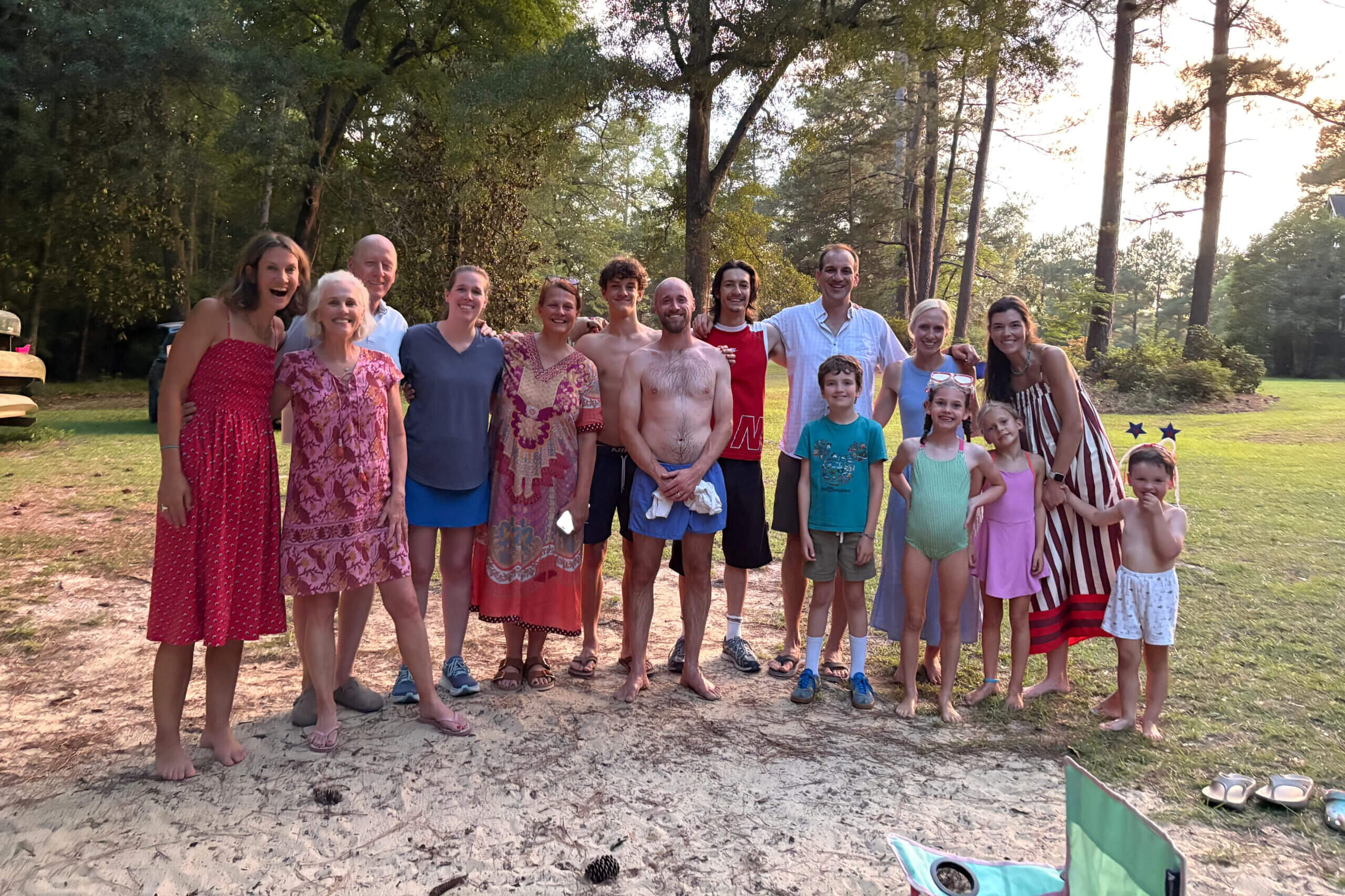 Group of adults and children standing arm-in-arm outdoors, smiling at a sunny park gathering with trees in the background.