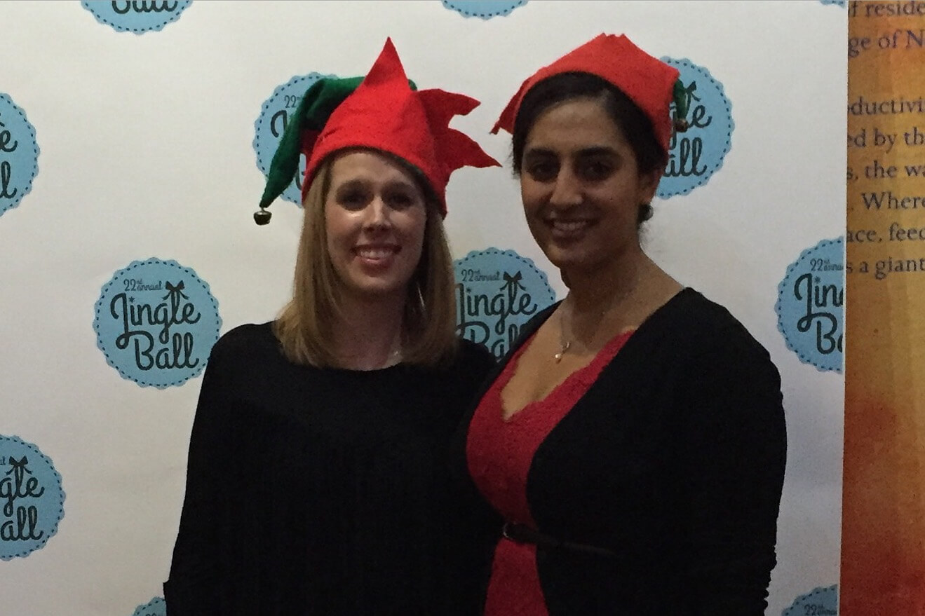 Two women pose for a photo at a Jingle Ball event, both wearing festive red elf hats. One in a black top, the other in a red dress with a black cardigan, standing in front of a branded backdrop.