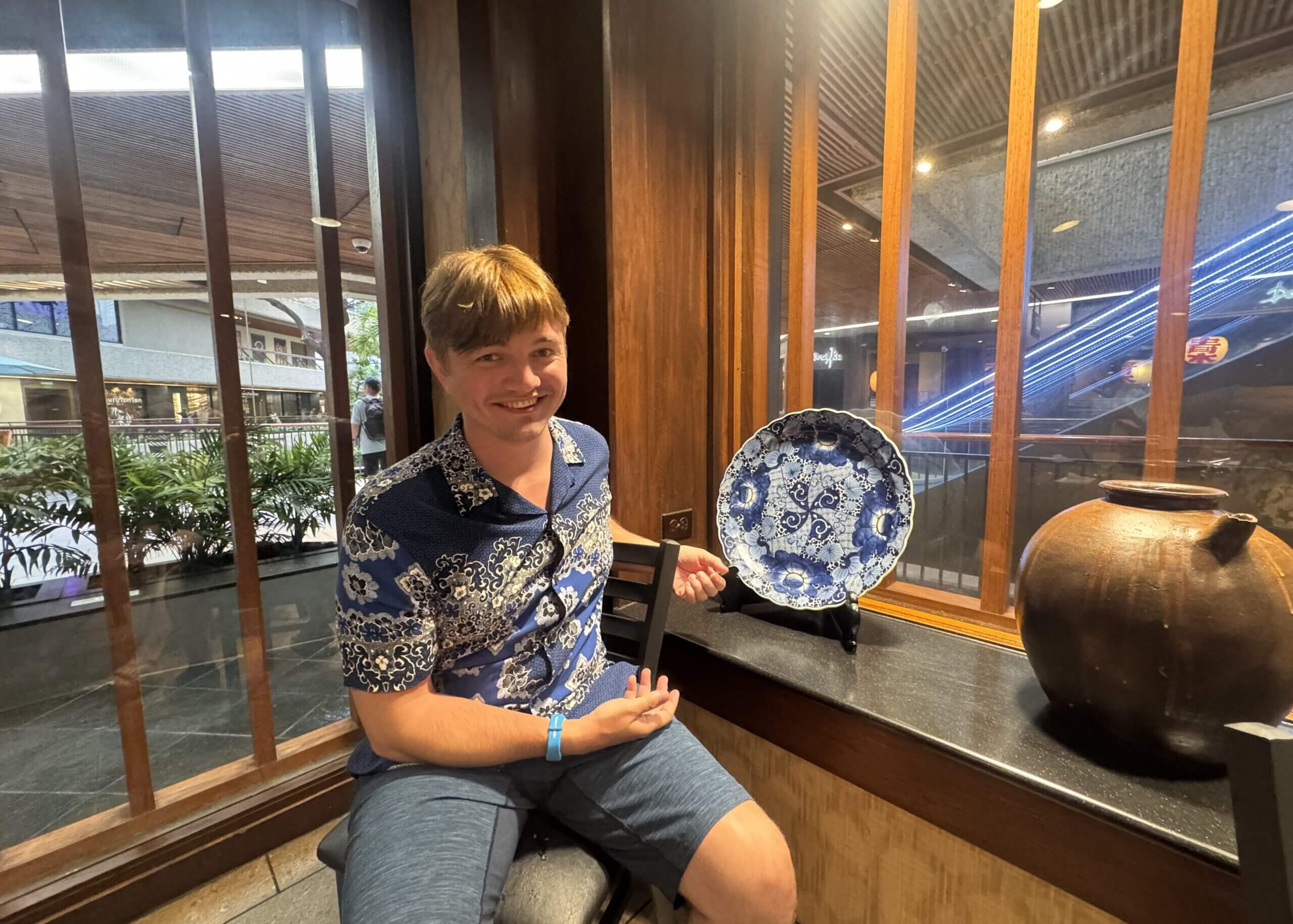 Smiling man in a blue floral shirt sits beside a blue-and-white patterned porcelain plate inside a wood-paneled display area.