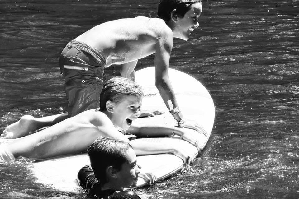Three kids ride a large inflatable board on a shimmering body of water; one stands at the back while two lie forward, smiling and holding on.