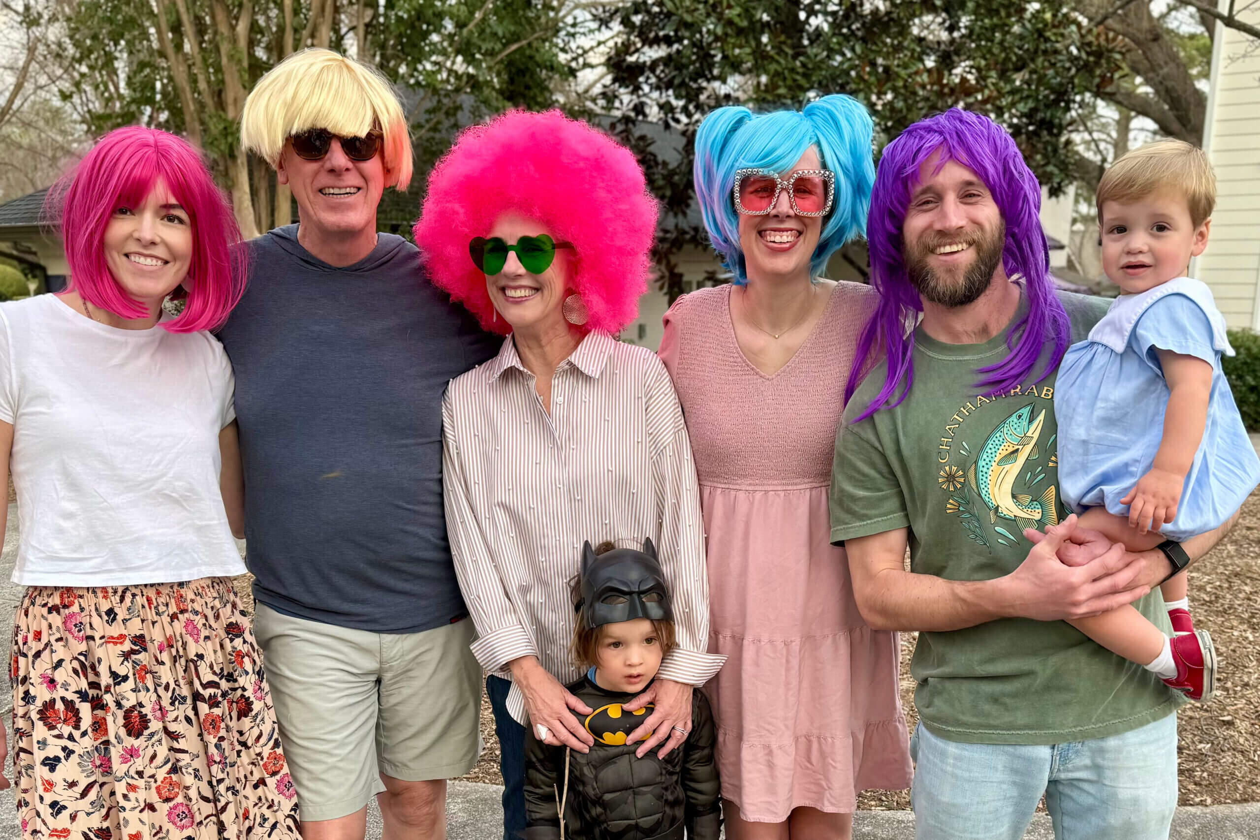 Group of adults and kids posing outdoors in bright, goofy wigs and accessories, smiling at the camera.