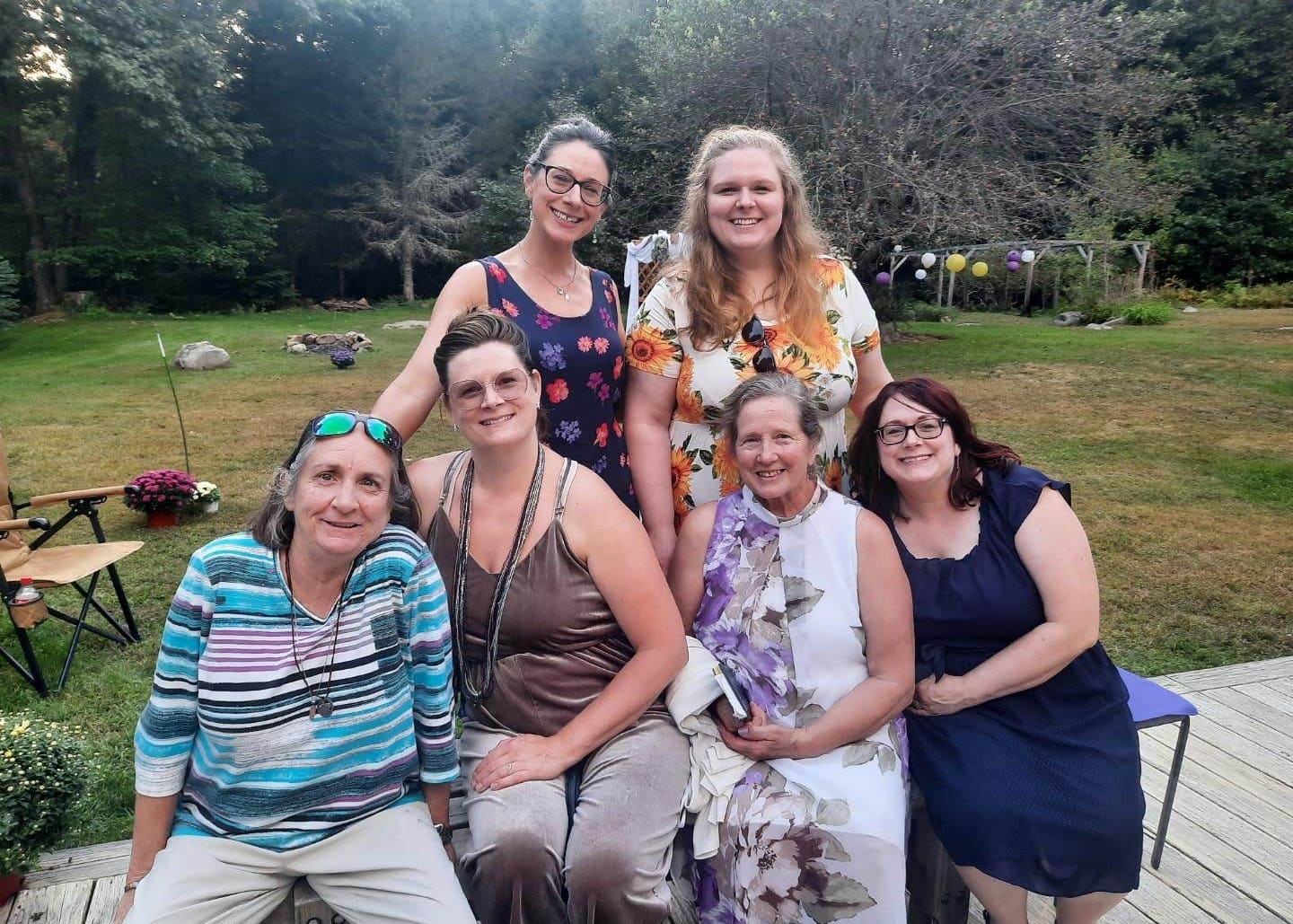 Group of six women smiling, posing outdoors on a wooden deck with a grassy yard and trees behind them; summer gathering setting with balloons in the background.
