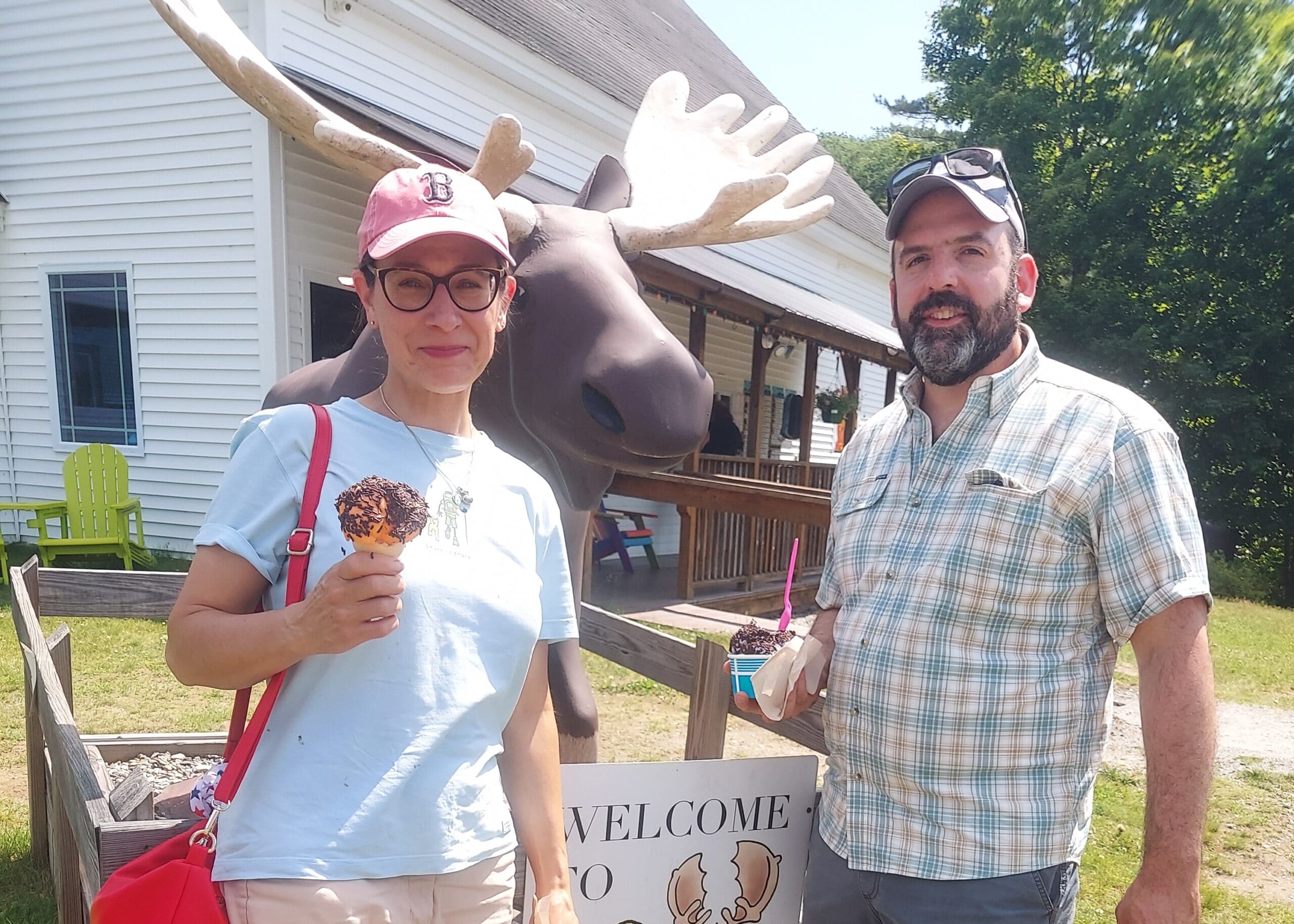 Two adults pose with a large moose statue and ice cream cones outdoors at a roadside attraction.