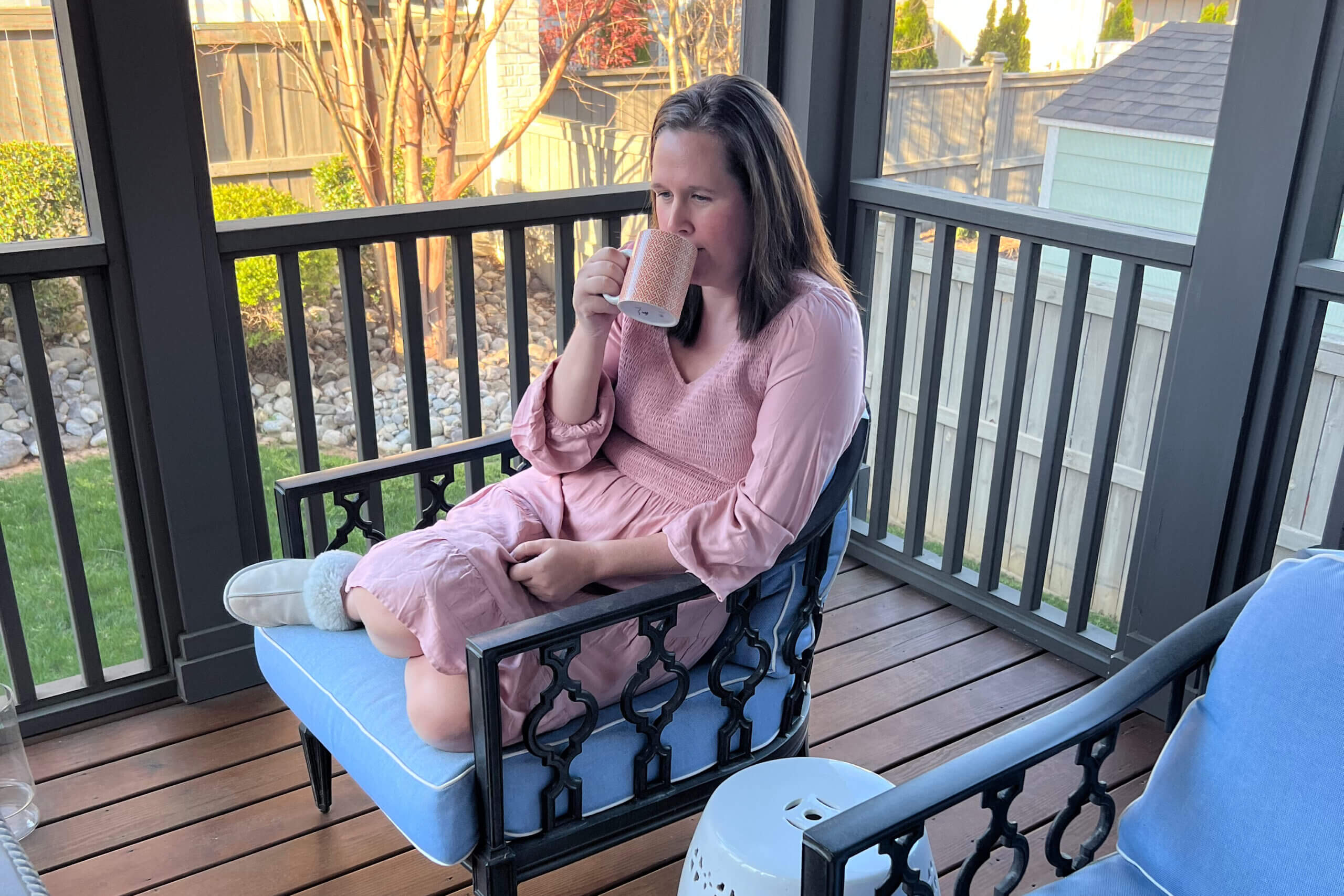 Woman in a pink dress relaxing on a blue-cushioned chair on a wooden porch, sipping from a patterned mug.