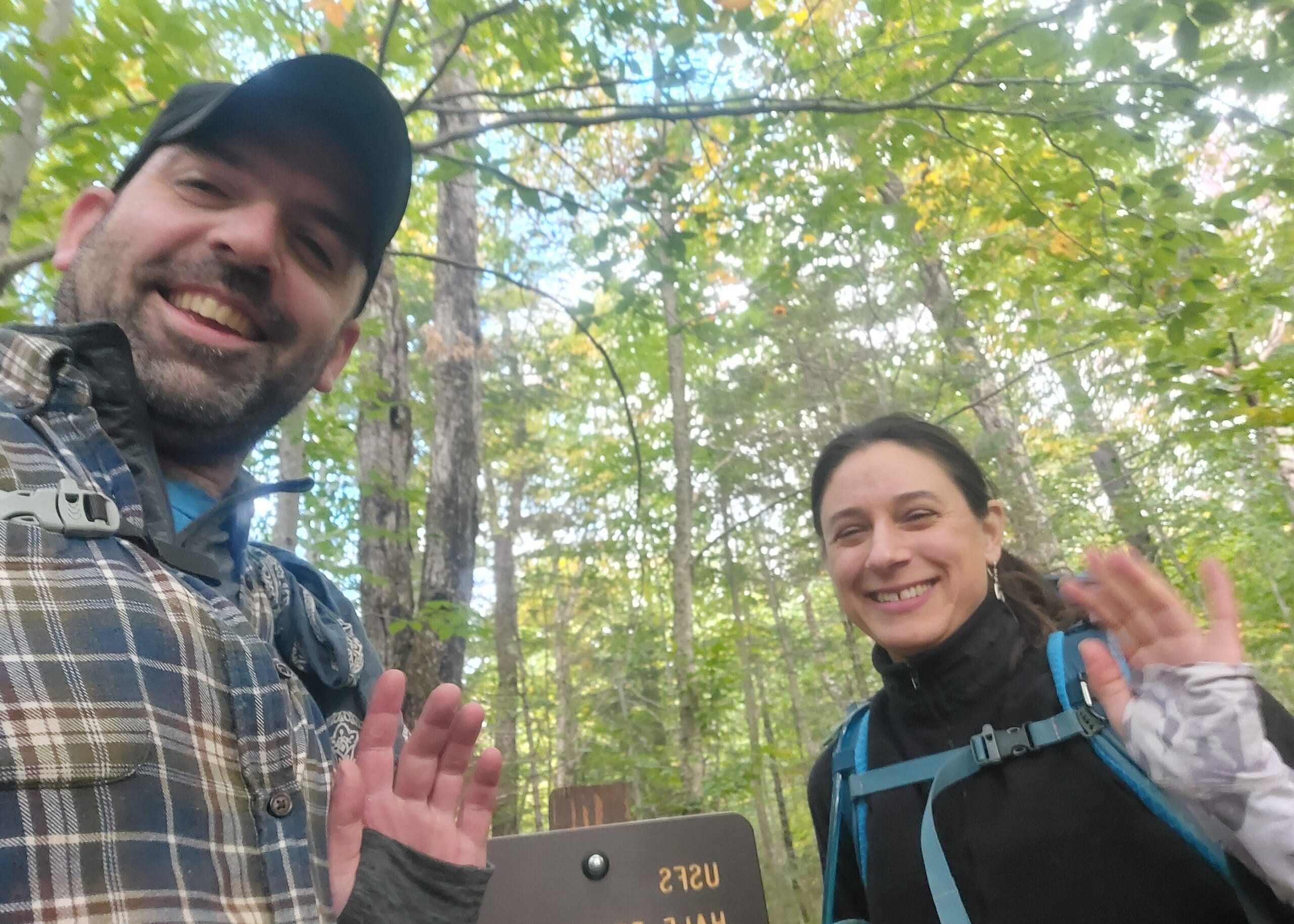 Two hikers smiling and waving at the camera in a leafy forest; man on the left wearing a plaid shirt and cap, woman on the right in hiking gear.