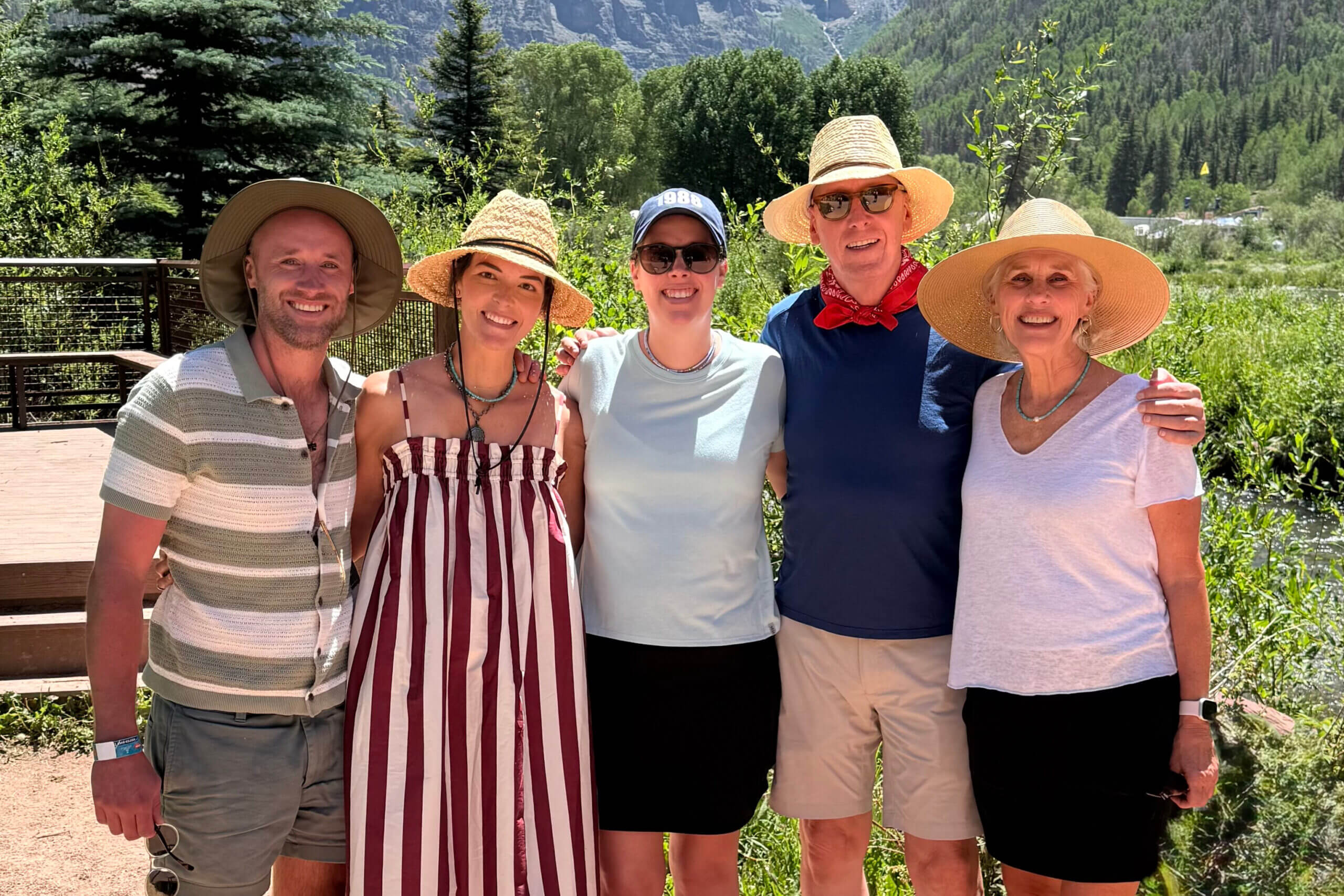 Five adults posing together outdoors on a sunny day, wearing hats, with trees and mountains in the background.
