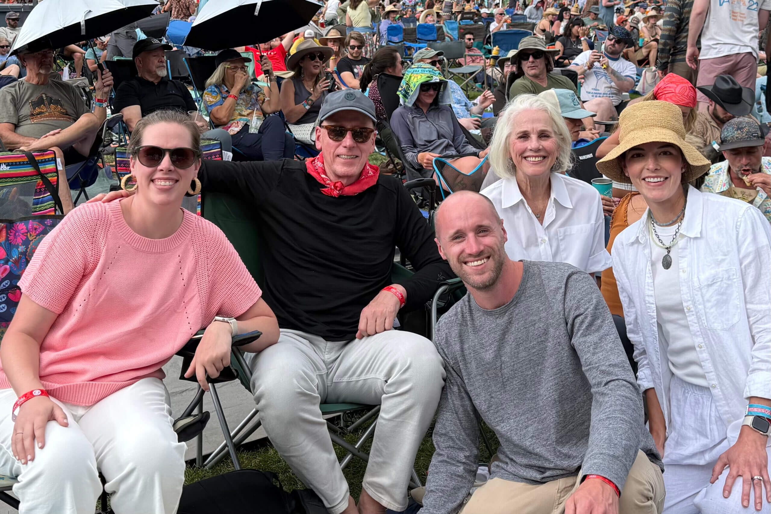 Group of smiling adults seated in folding chairs at an outdoor festival with a crowded crowd behind them.