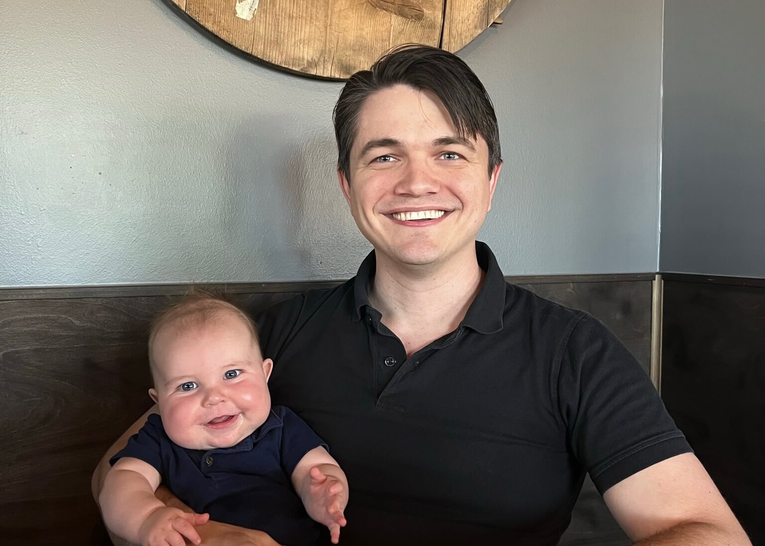 Smiling man in a black polo holds a cheerful baby in a navy shirt, seated in a booth against a blue wall.