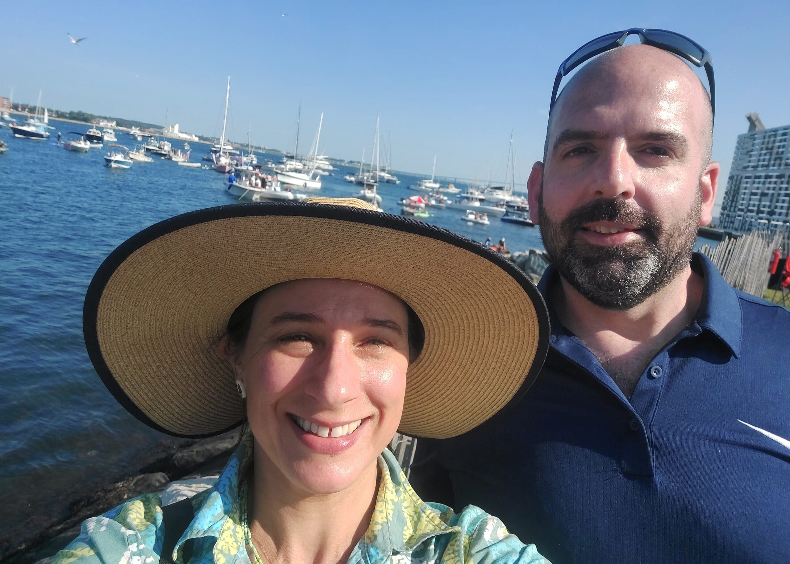 Smiling couple taking a seaside selfie with a harbor full of boats behind them on a sunny day, wearing hats and sunglasses.