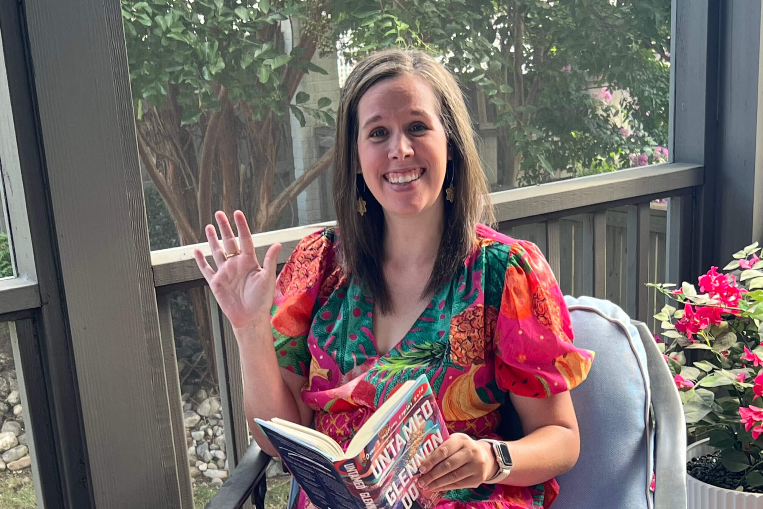 Smiling woman in a bright tropical dress sits on a porch, waves at the camera, and holds an open magazine.
