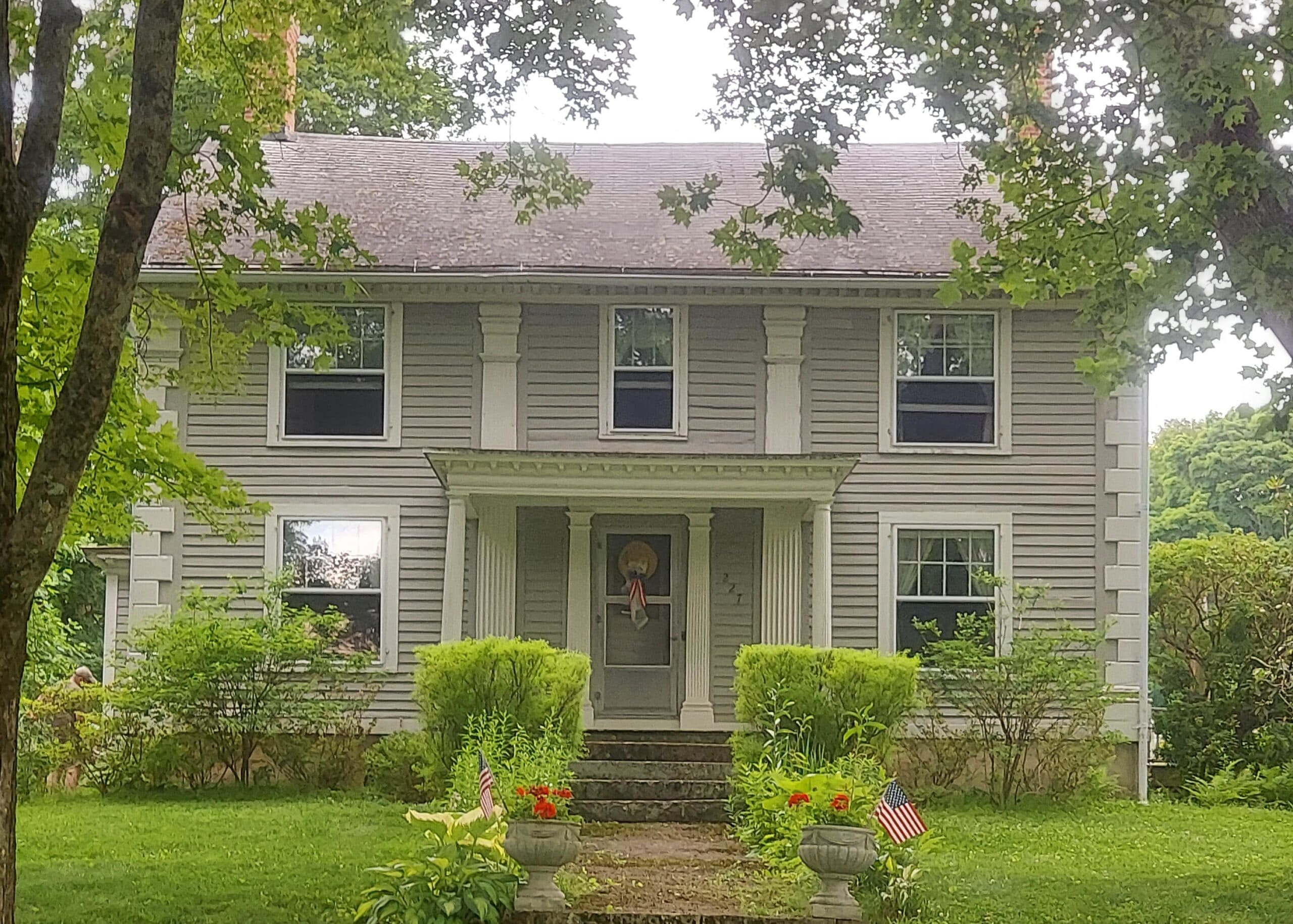 Front view of a gray two-story house with a covered porch, flanked by trees and a landscaped yard with small American flags in planters.