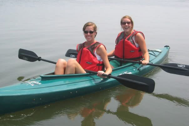 Two people in a teal tandem kayak wearing red life jackets paddle on calm water, smiling at the camera.