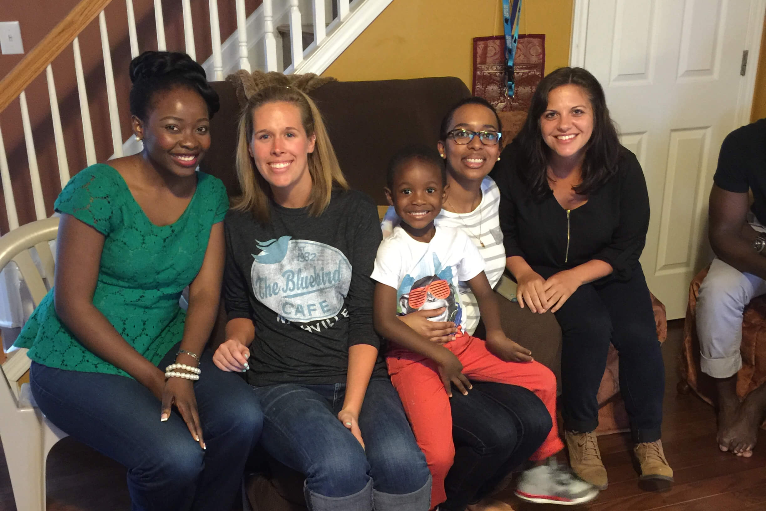 Group of six people (five adults and a child) sitting on a couch indoors, smiling at the camera.