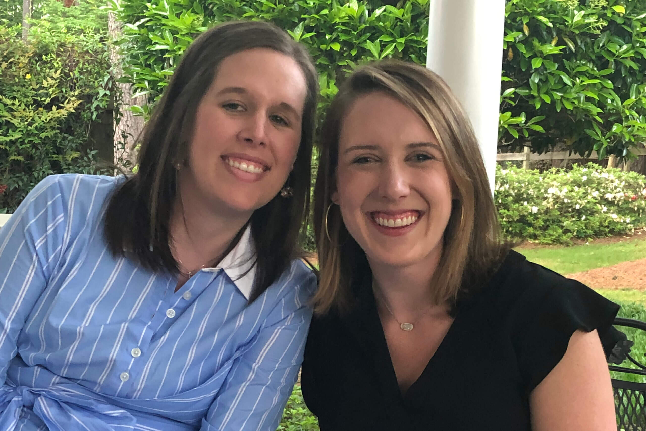 Two women sit closely together on a bench outdoors, smiling at the camera with green bushes in the background (one in a blue striped shirt, the other in black).