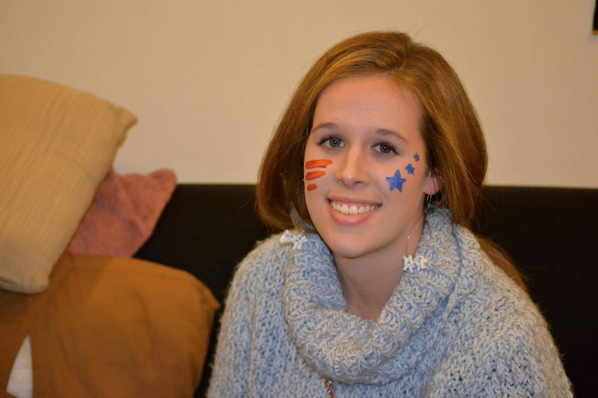 Smiling girl with blue star face paint on her right cheek and orange stripes on her left cheek, wearing a chunky blue sweater.