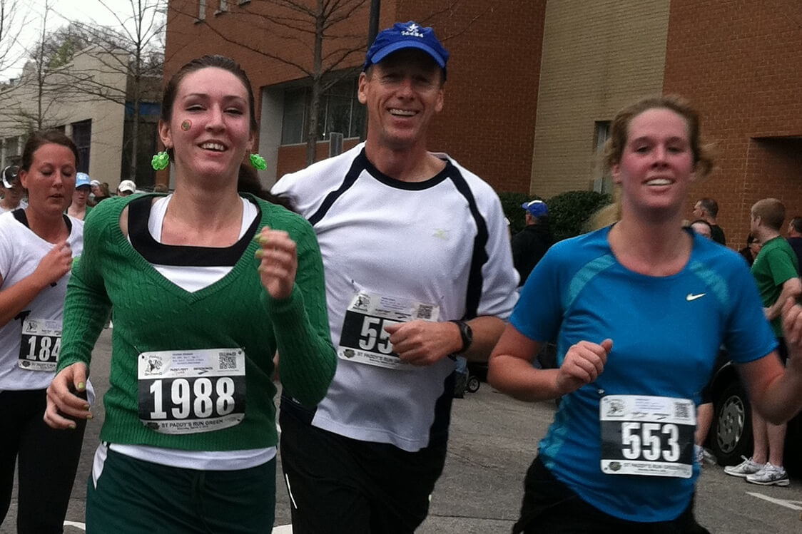 Smiling group of runners in a street race; bibs 1988 and 553 visible. A woman in green sweater leads, flanked by a man in white and a woman in blue.