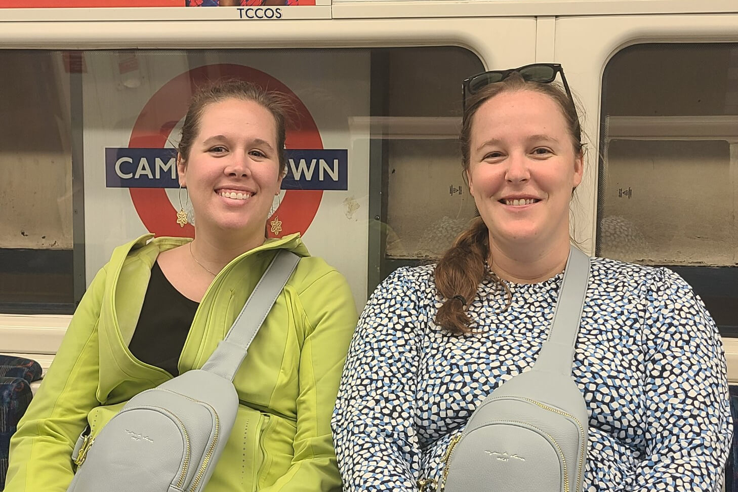 Two smiling women sitting on a London Underground train with a Camden Town sign behind them, both wearing light gray crossbody bags.